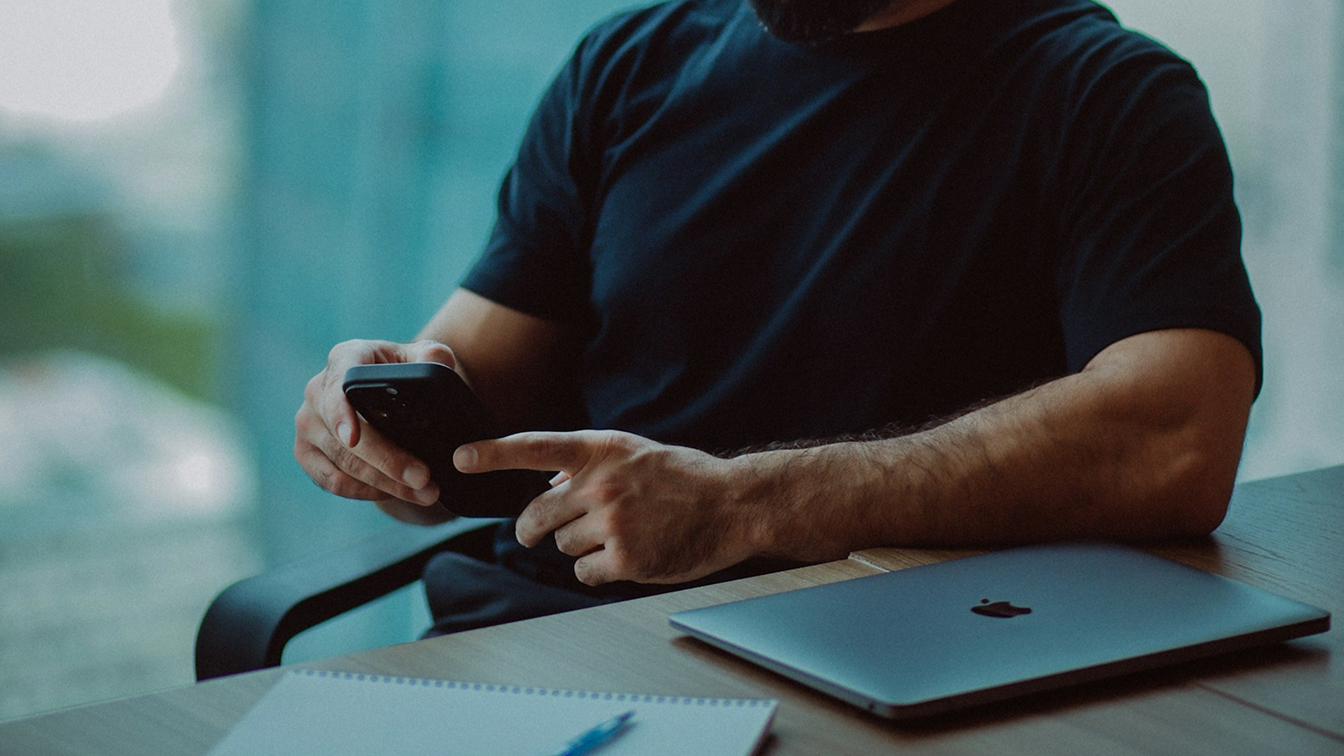 Person sitting at a desk using a smartphone next to a laptop, representing interaction with digital systems and workflows. 