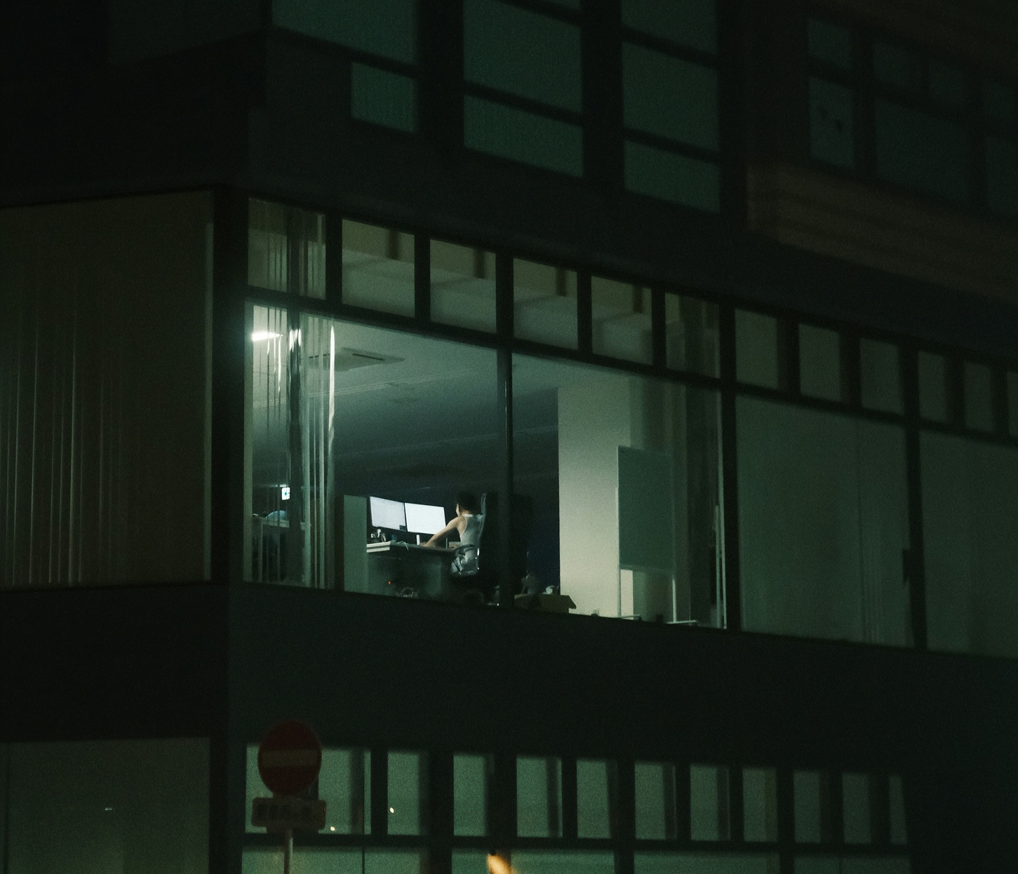 Lone employee at dual monitors in a lit office window at night, a sign of the off-hours activity that signals insider risk