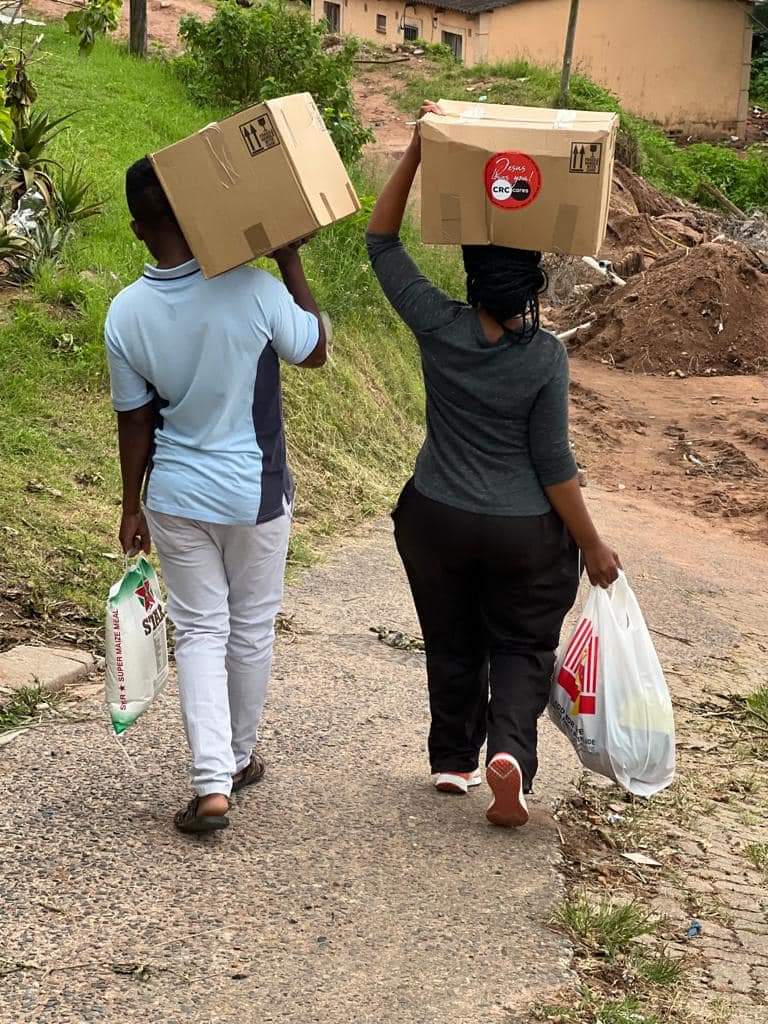 Two people walking on a paved path carrying cardboard boxes on their heads and plastic bags in their hands.