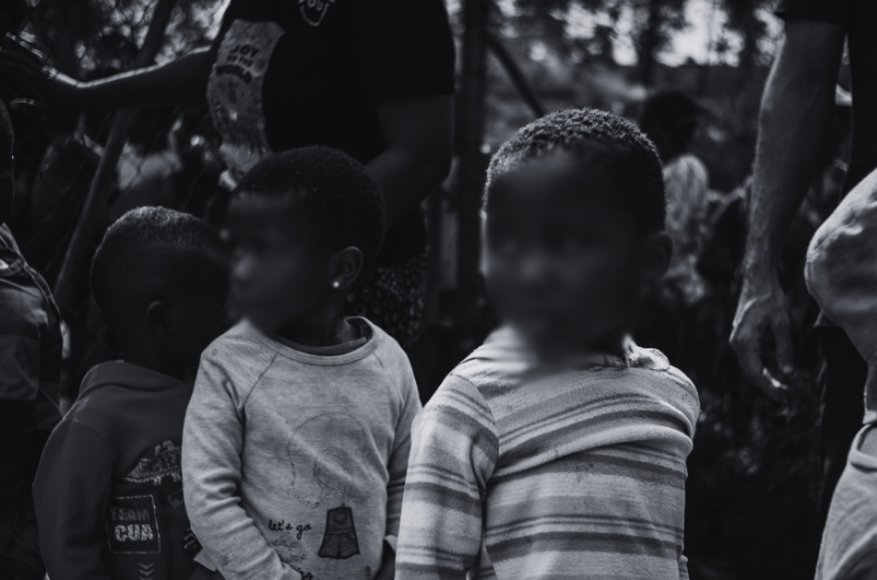 Black and white photo of young children standing outdoors, some adults partially visible in background.