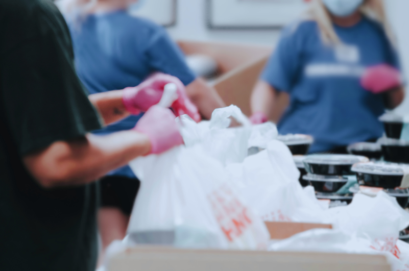 Volunteers wearing gloves packing food containers and white plastic bags on a table.