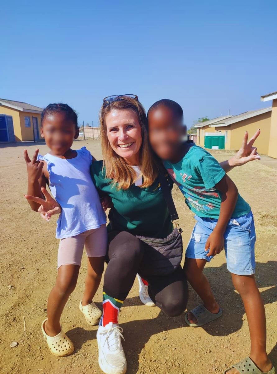 Woman kneeling outdoors with two children making peace signs, smiling under a clear blue sky.