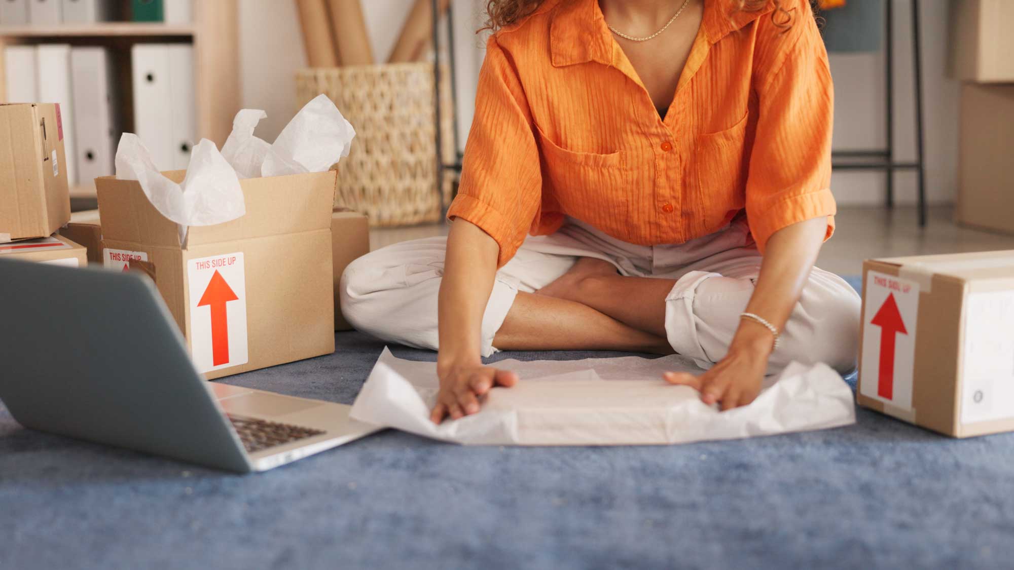 Person sitting on the floor, wrapping items with paper near boxes and a laptop.