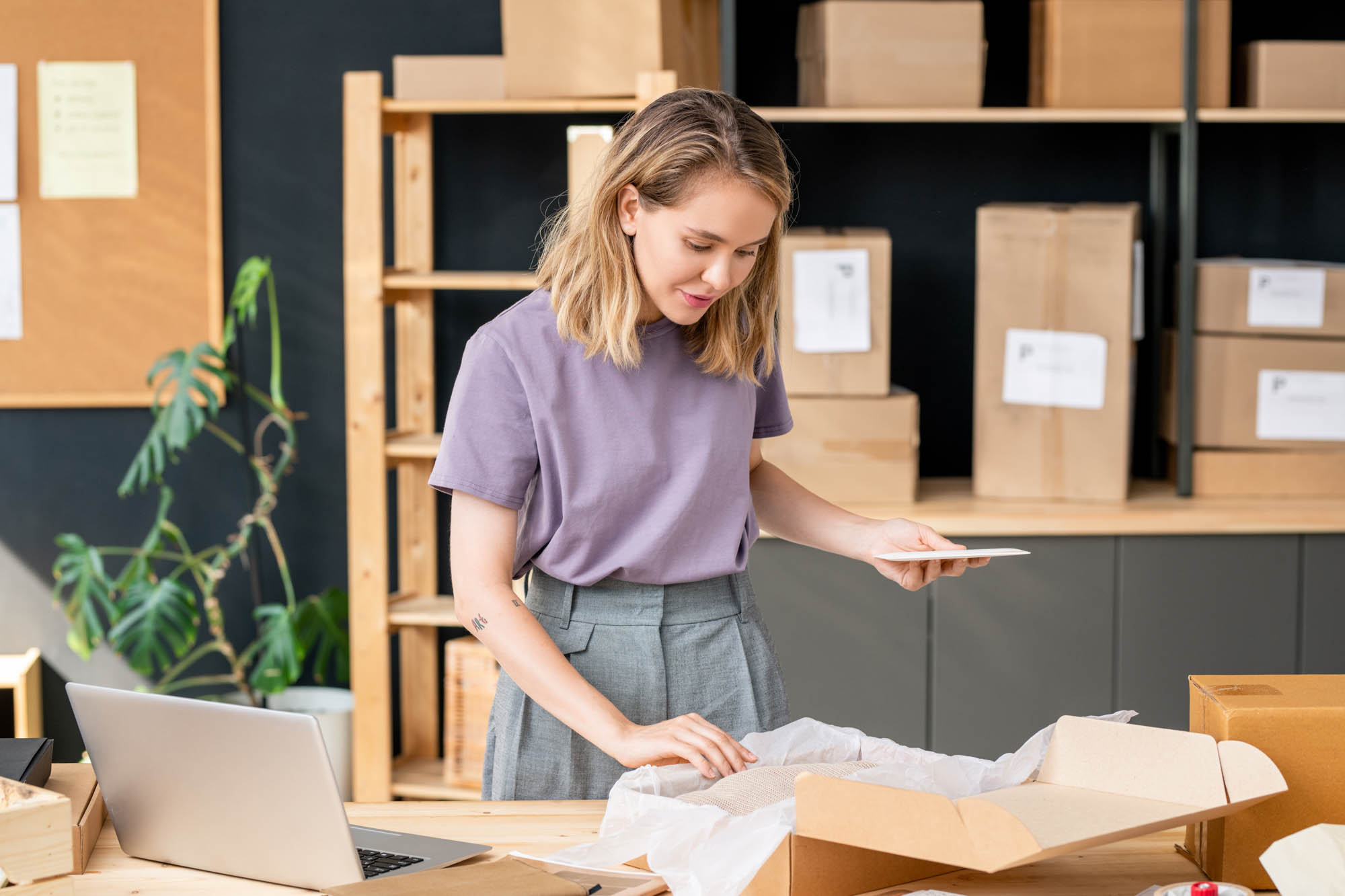 A woman stands at a table, packing a box with tissue paper in a workspace.