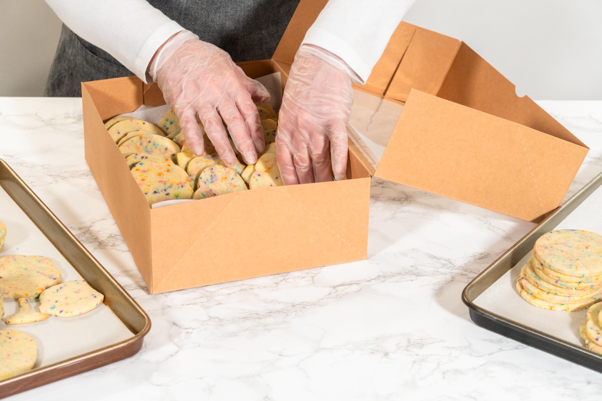 A person wearing gloves places sprinkle cookies into a brown box on a counter.