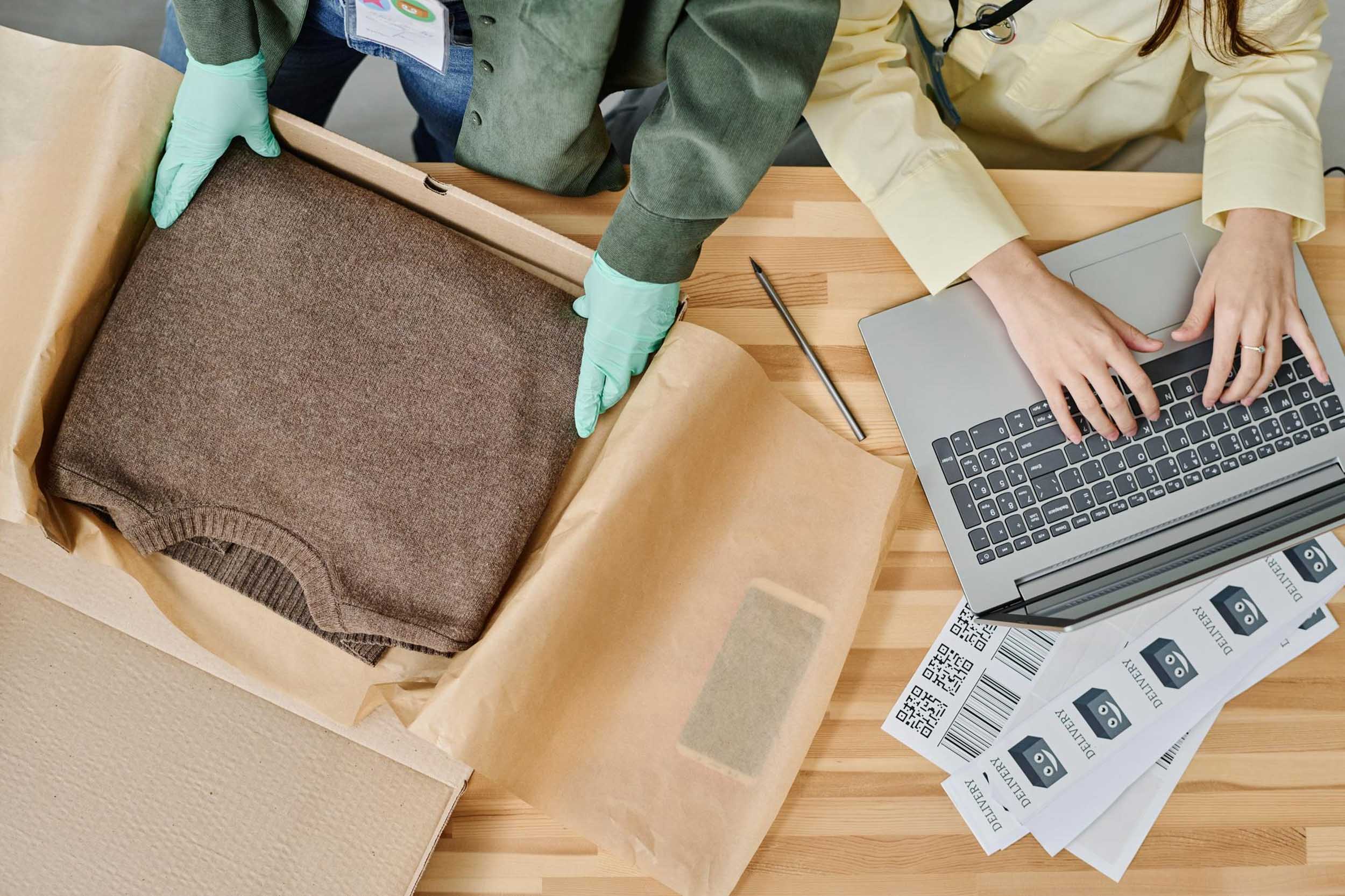 Person wearing gloves packs a brown sweater in a box, while another types on a laptop nearby.