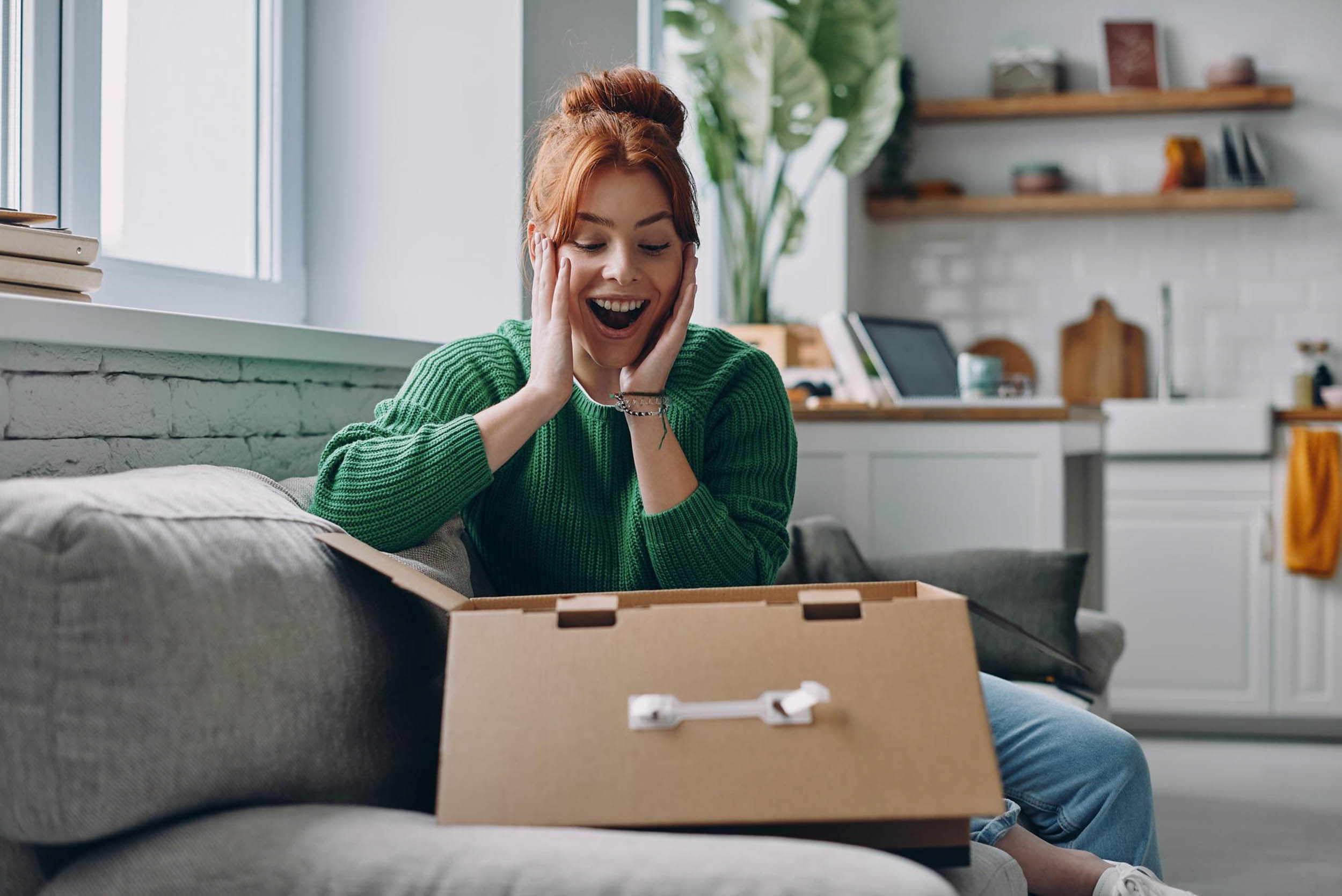 A woman looks surprised while opening a cardboard box on a couch at home.