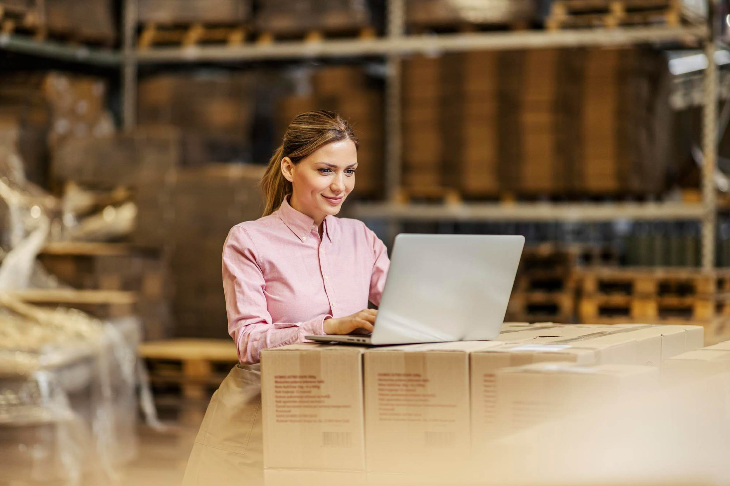 Woman in pink shirt using a laptop while standing by stacked cardboard boxes in a warehouse.