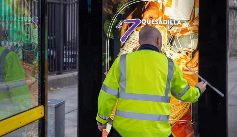 White man wearing a hi-vis jacket, cleaning a bus shelter advertising screen