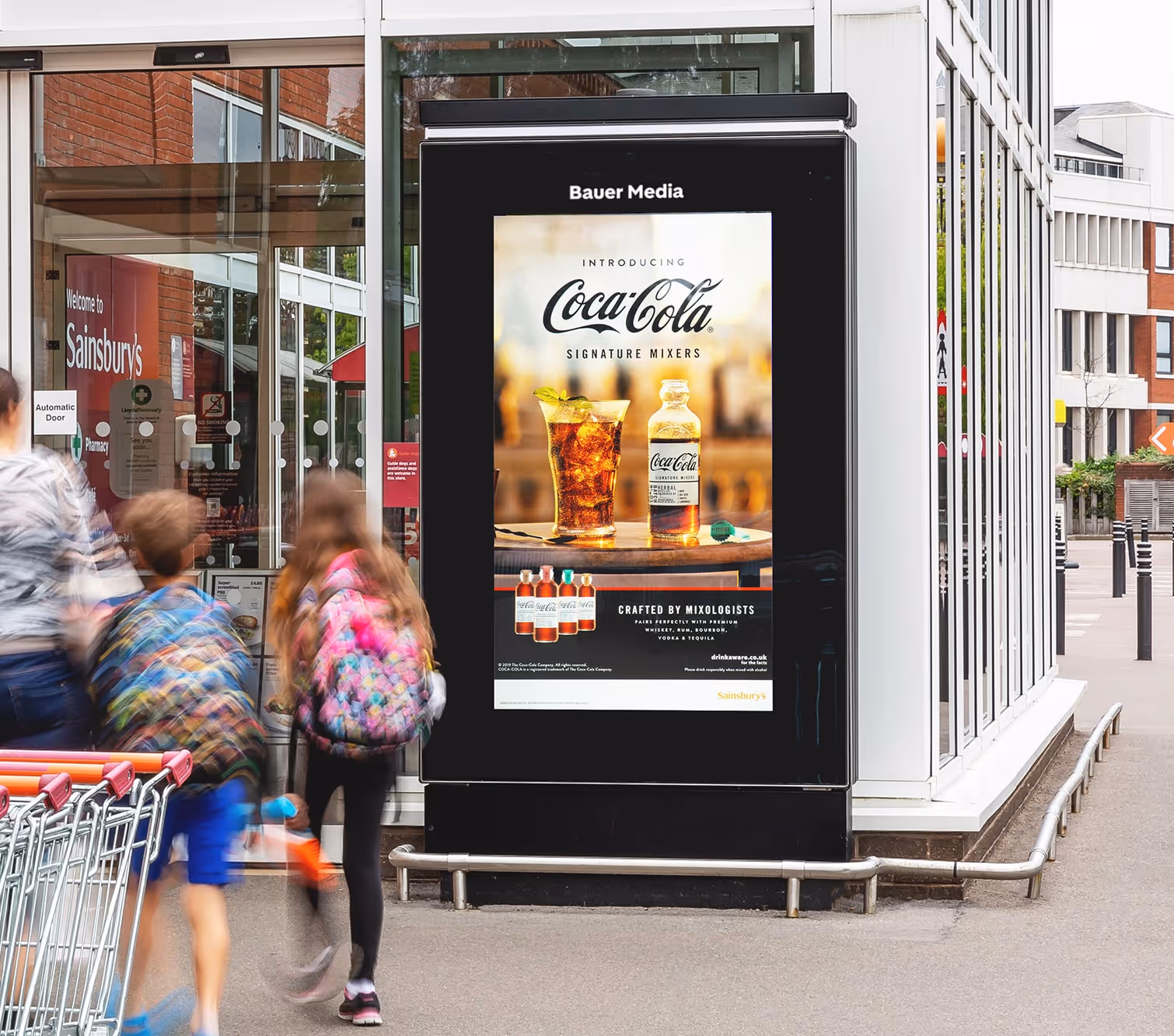 A Coco Cola advert on a supermarket digital screen with people walking into the store