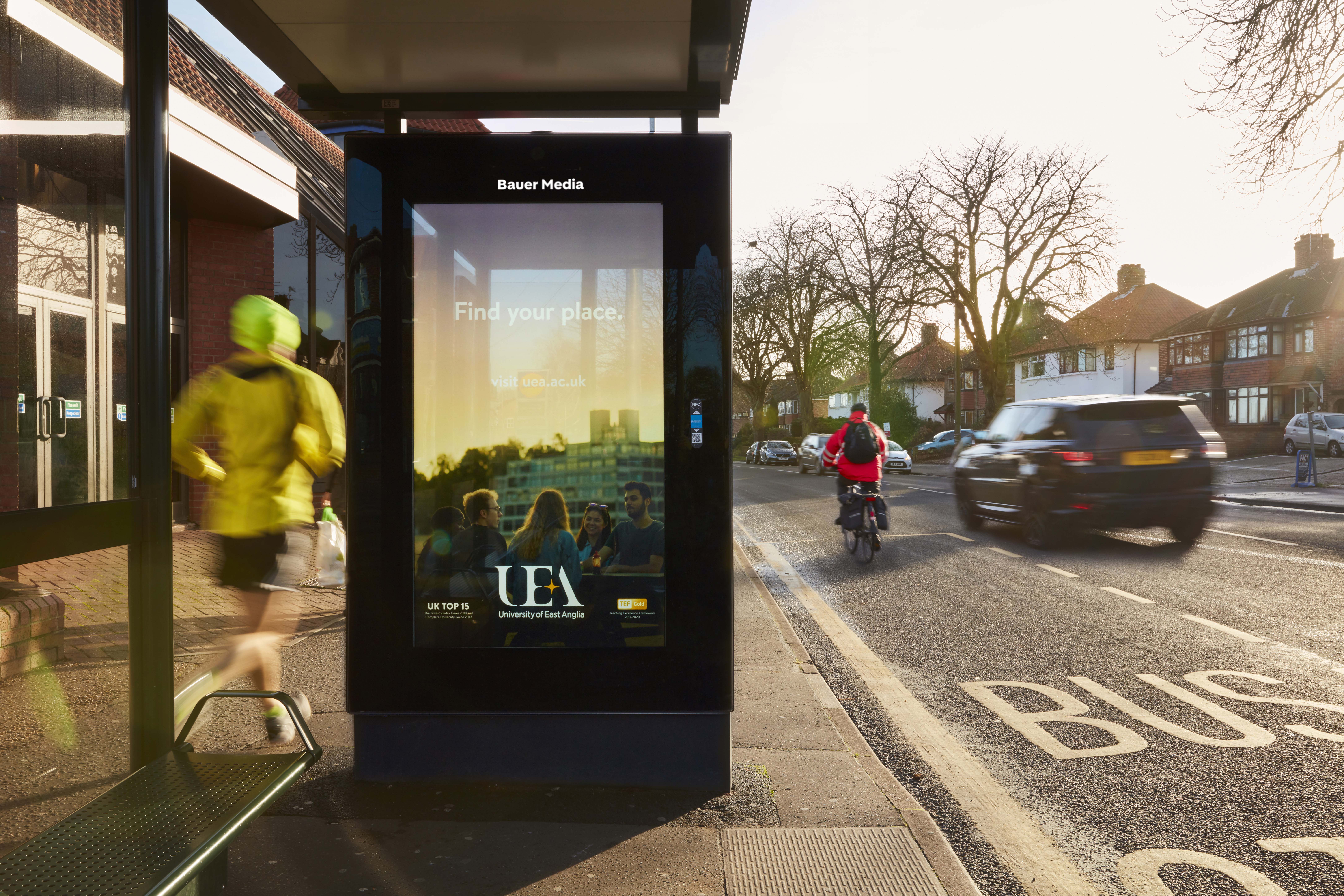 UEA's Bus Shelter ad as people run and cycle past