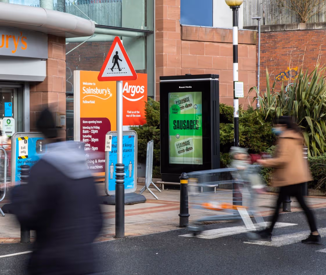 A digital Sainsbury's advertising screen at the entrance displaying a sausages campaign