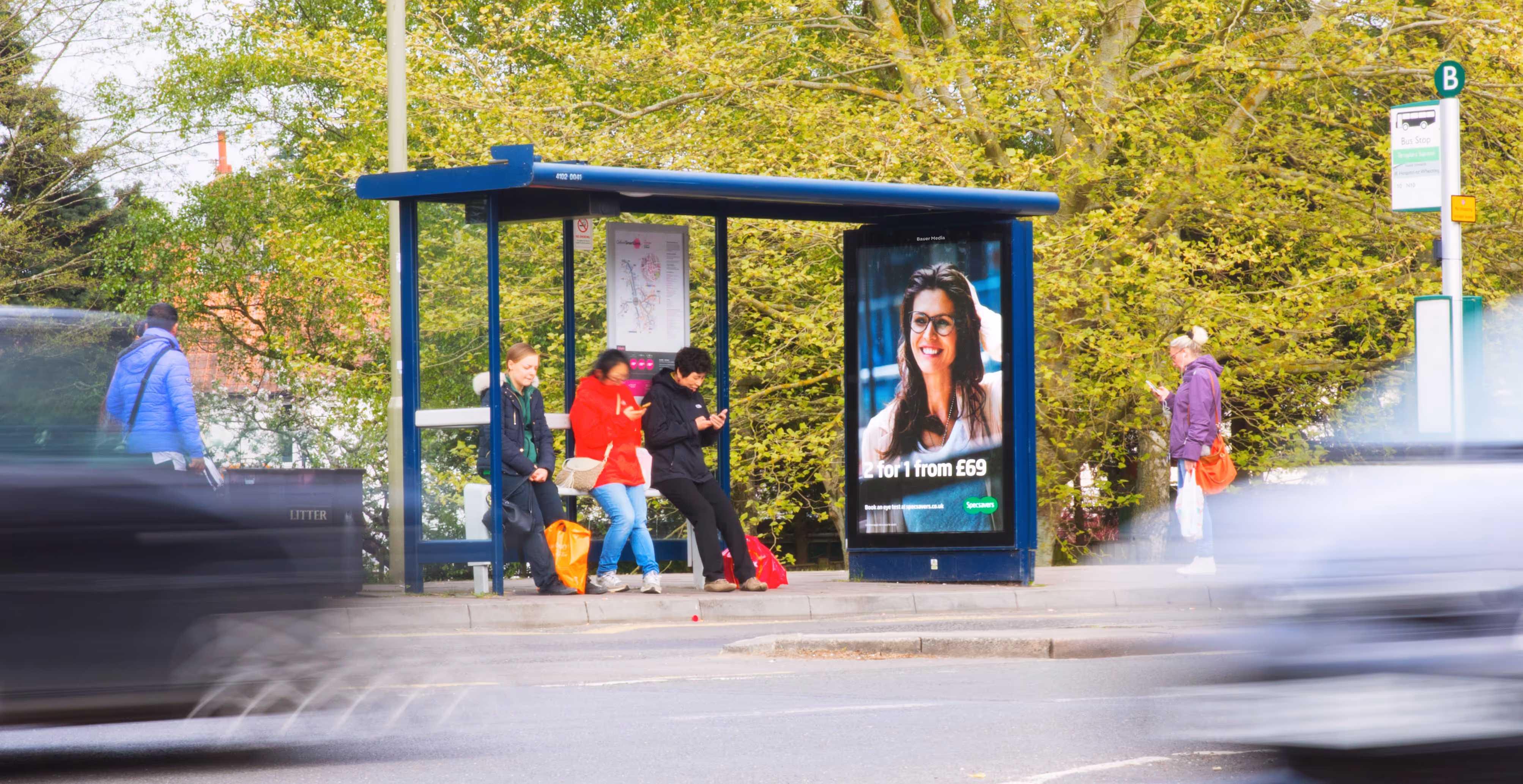 A Specsavers ad campaign displaying on a bus shelter poster with people sat on the bench