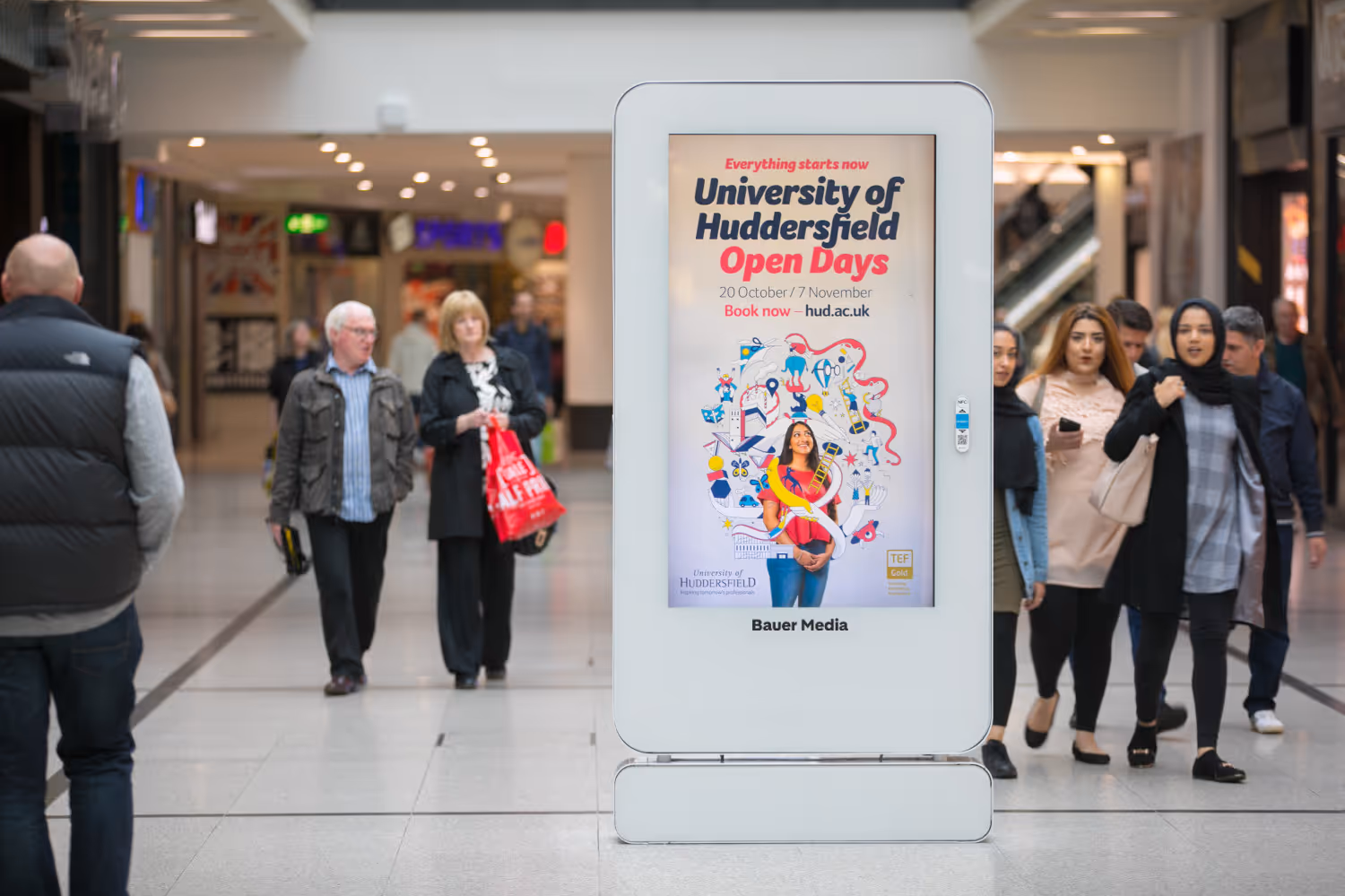 A Huddersfield Uni ad campaign displaying on a digital shopping centre screen with shoppers walking by