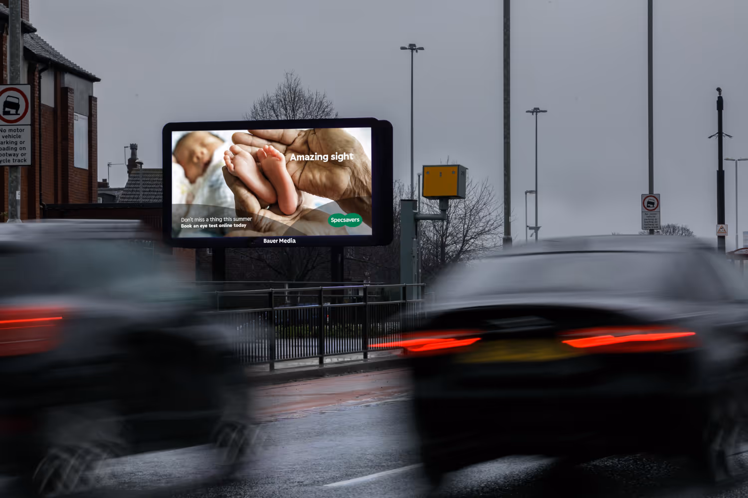 Bauer Media Outdoor Billboard digital advertising screen on a roadside displays a Specsavers ad featuring baby feet and the message "Amazing sight," as cars drive past.