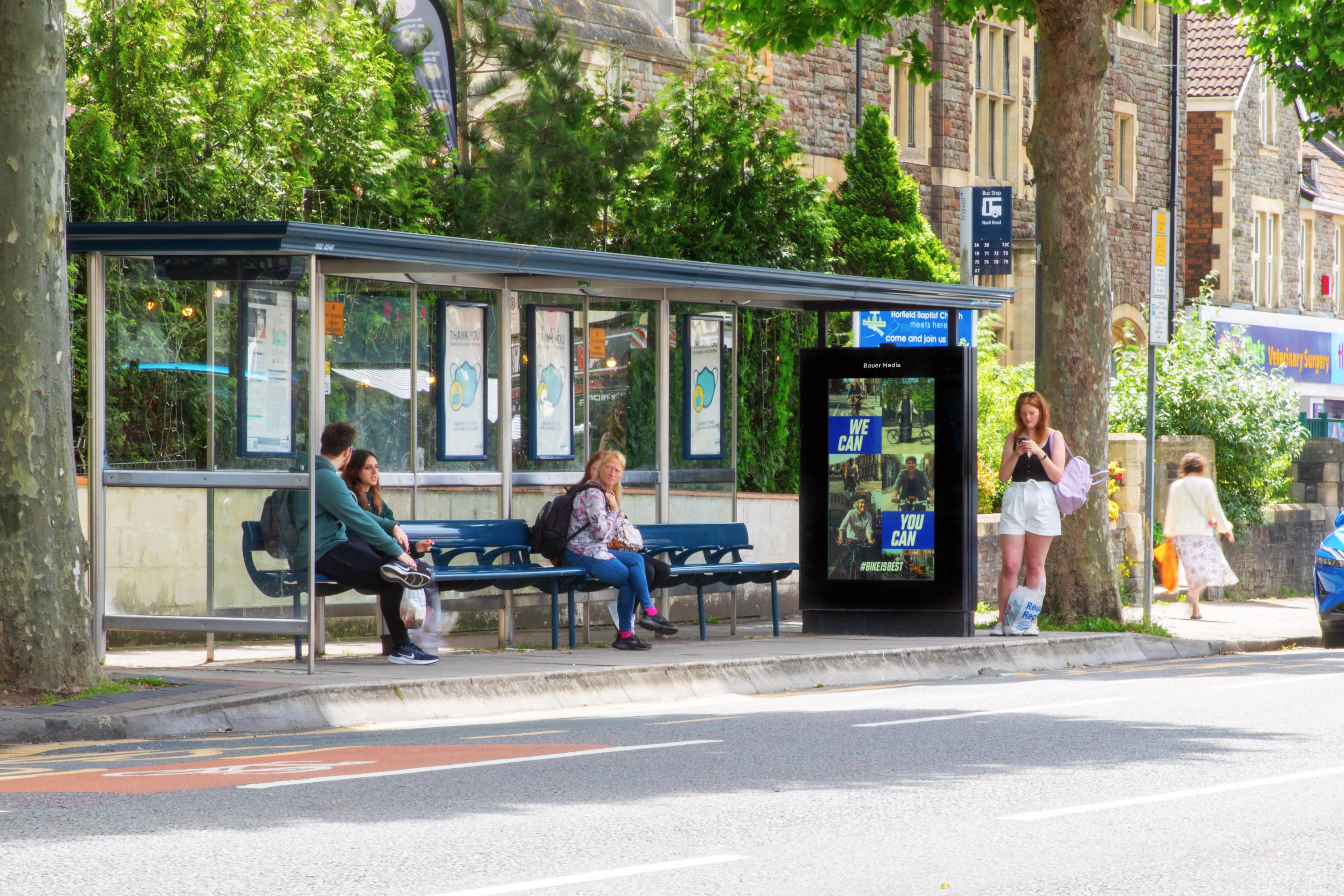 Busy bus shelter with people waiting for a bus while an advertisement is being shown on the bus shelter screen