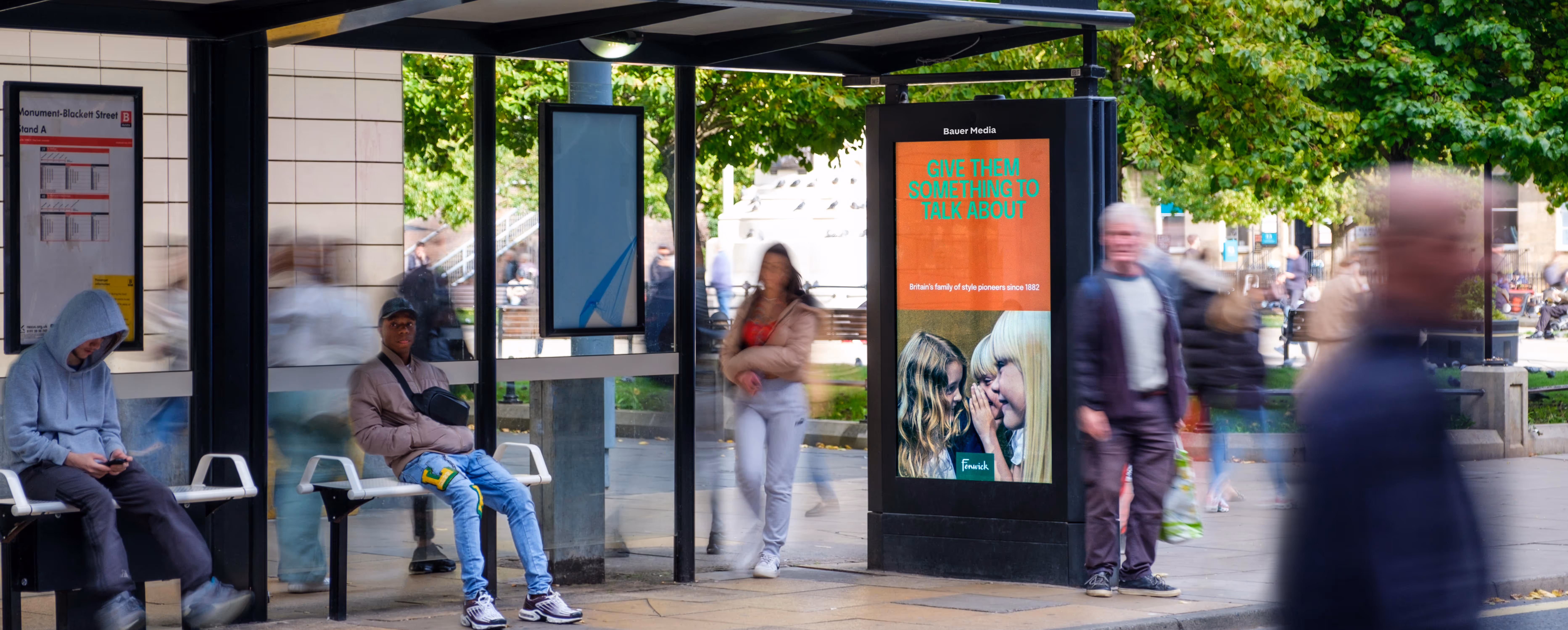 Busy bus shelter with people waiting for a bus while an advertisement is being shown on the bus shelter screen
