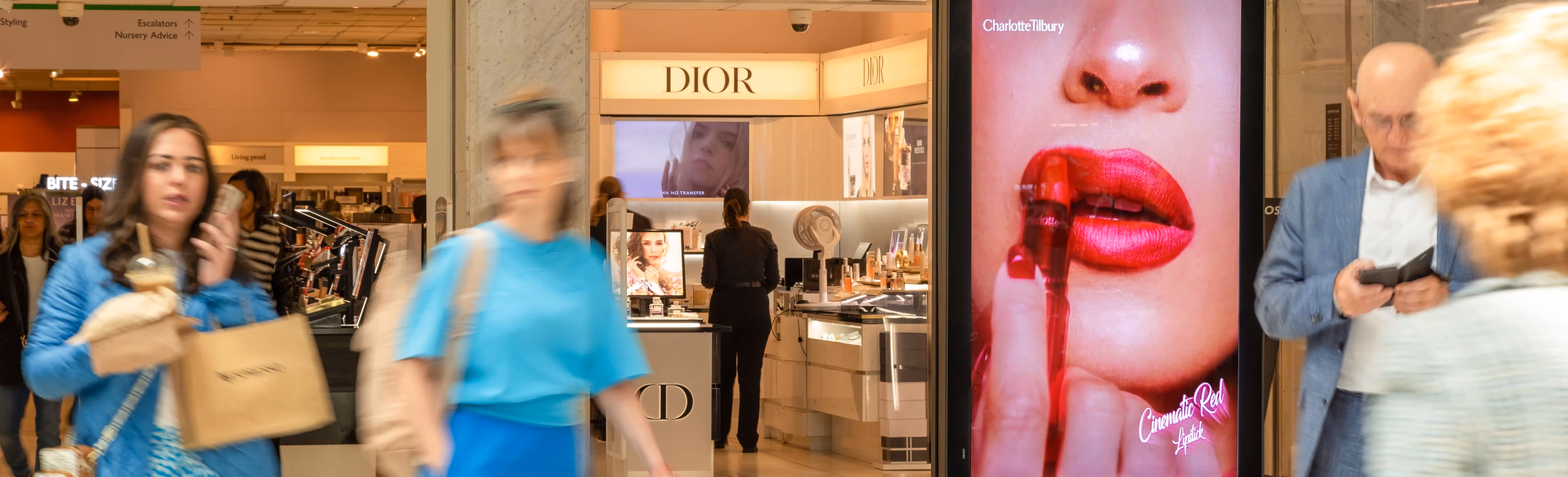 Bauer Media Outdoor Digital advertising screen in a malls displays a Charlotte Tilbury lipstick ad. Shoppers pass by, with the Dior beauty counter visible in the background.