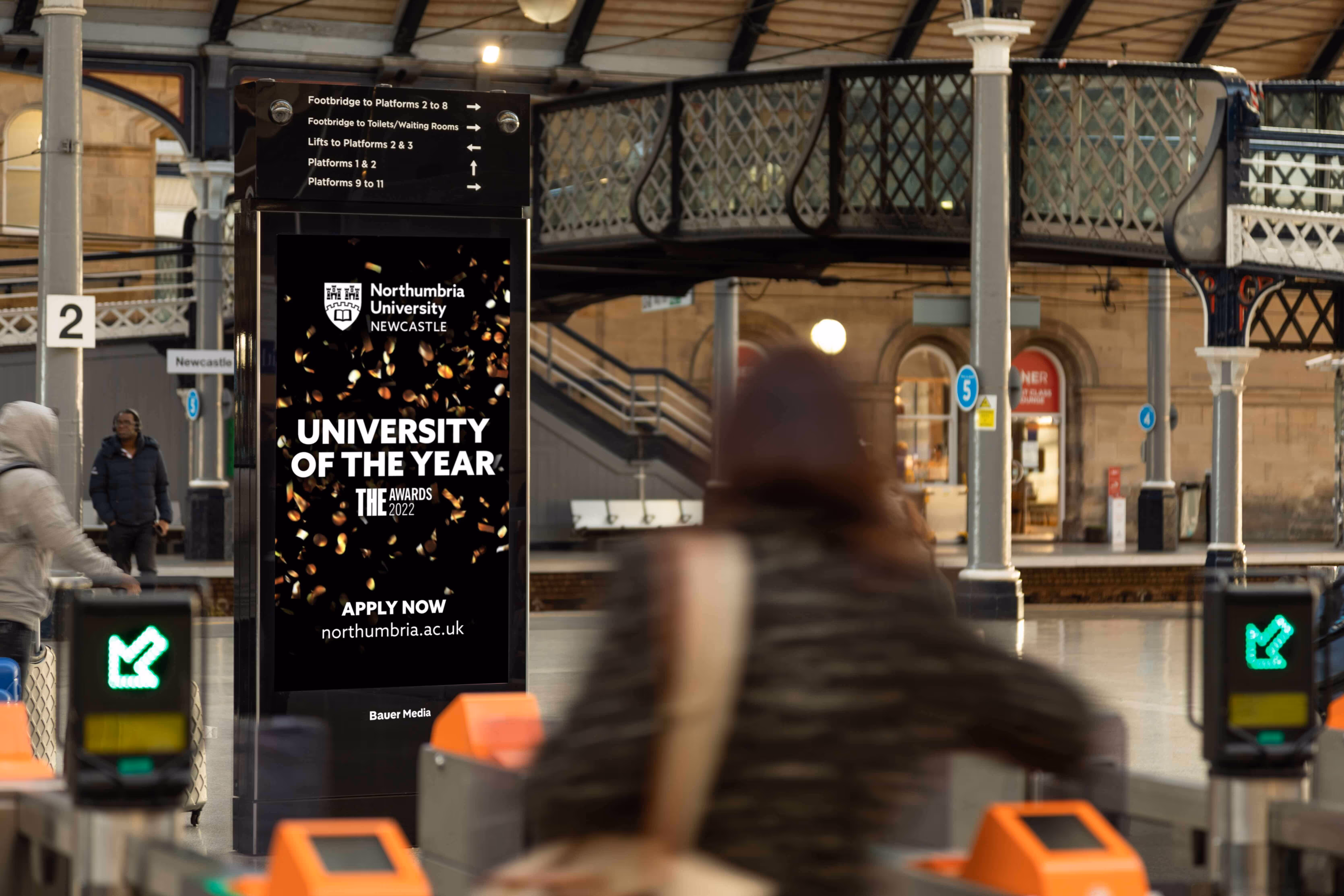 Bauer Media Outdoor digital advertising screen at a train station displays a Northumbria University ad promoting its “University of the Year” accolade, as commuters move through the concourse.