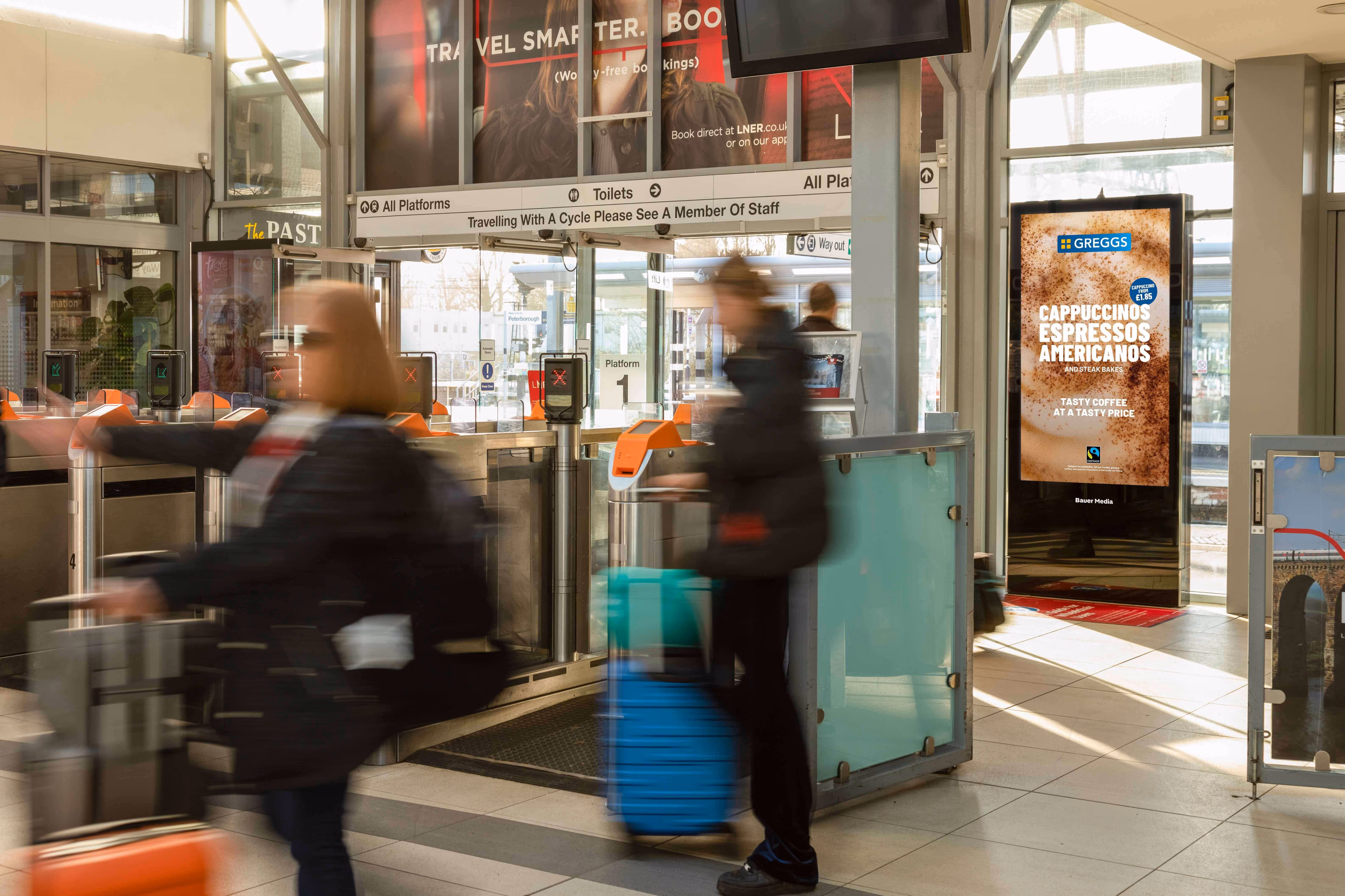 Bauer Media Outdoor digital advertising screen at a train station entrance displays a Greggs coffee ad, as commuters pass through ticket barriers.