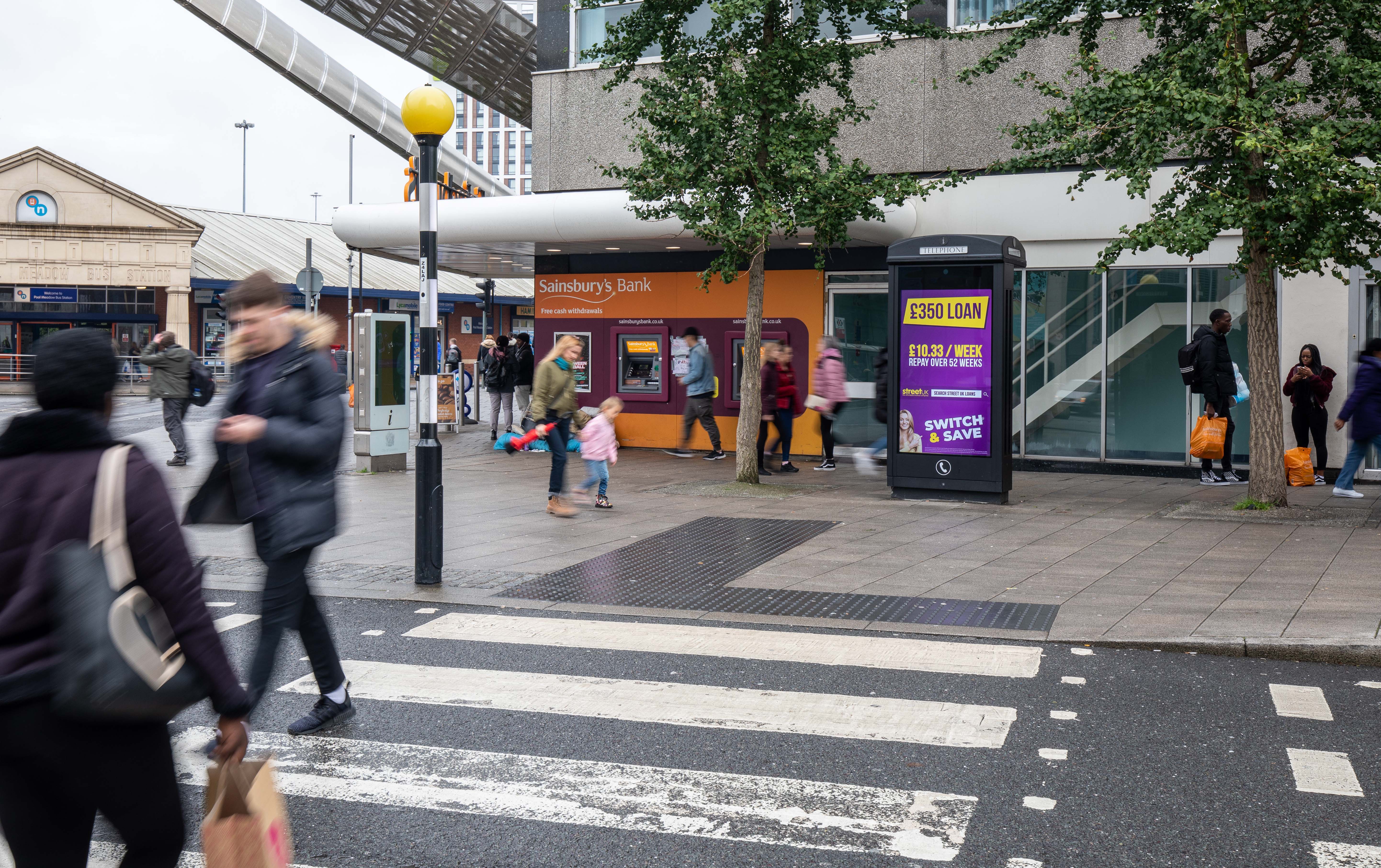 A digital payphone screen near a zebra crossing and outside of Sainsbury's