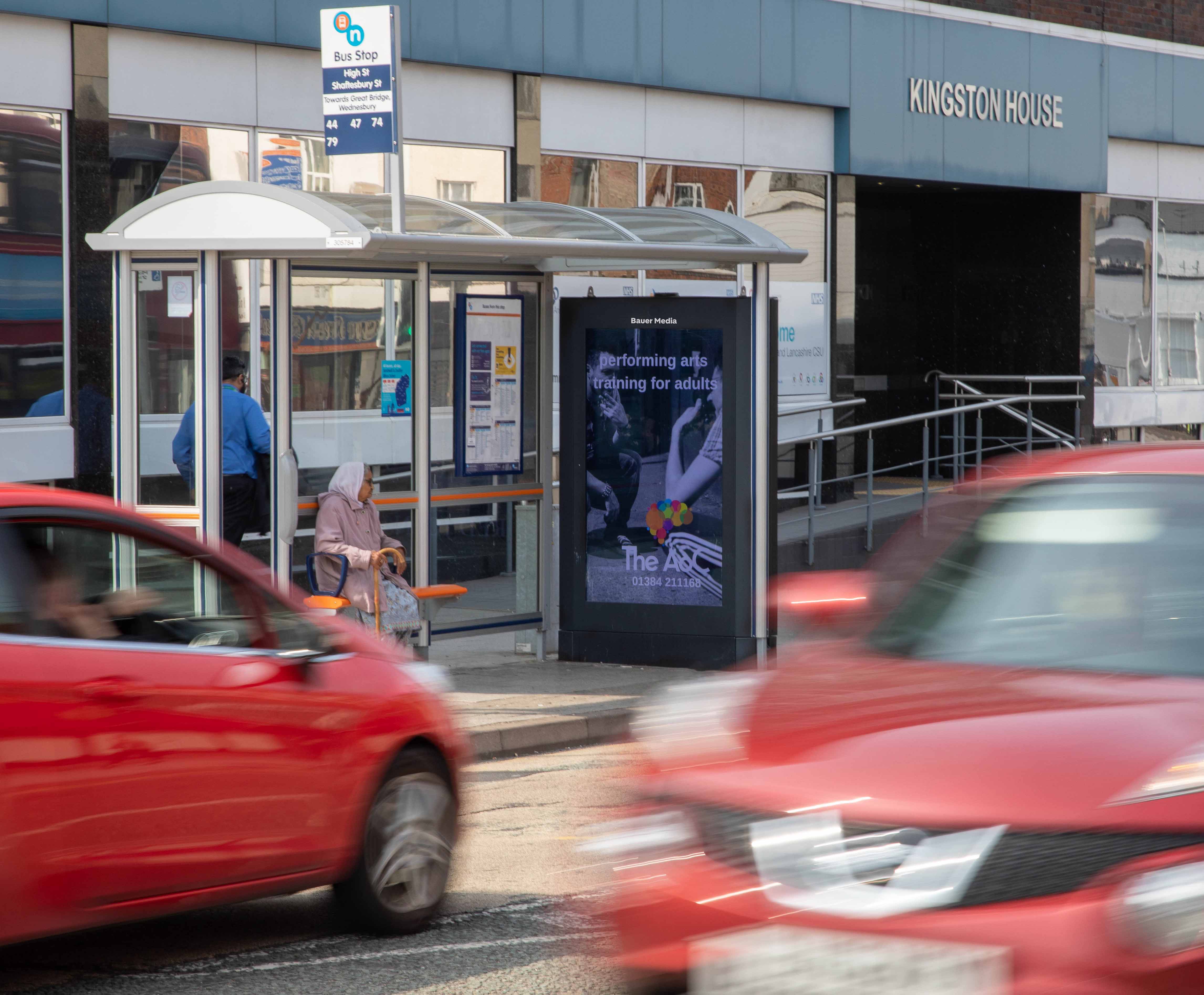 A digital bus shelter screen displaying a performing arts ad campaign with traffic driving past