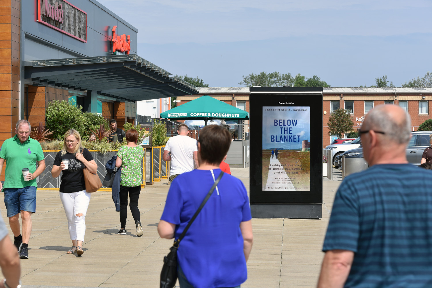 A digital advertising screen on a retail park, outside of restaurants, with people walking by