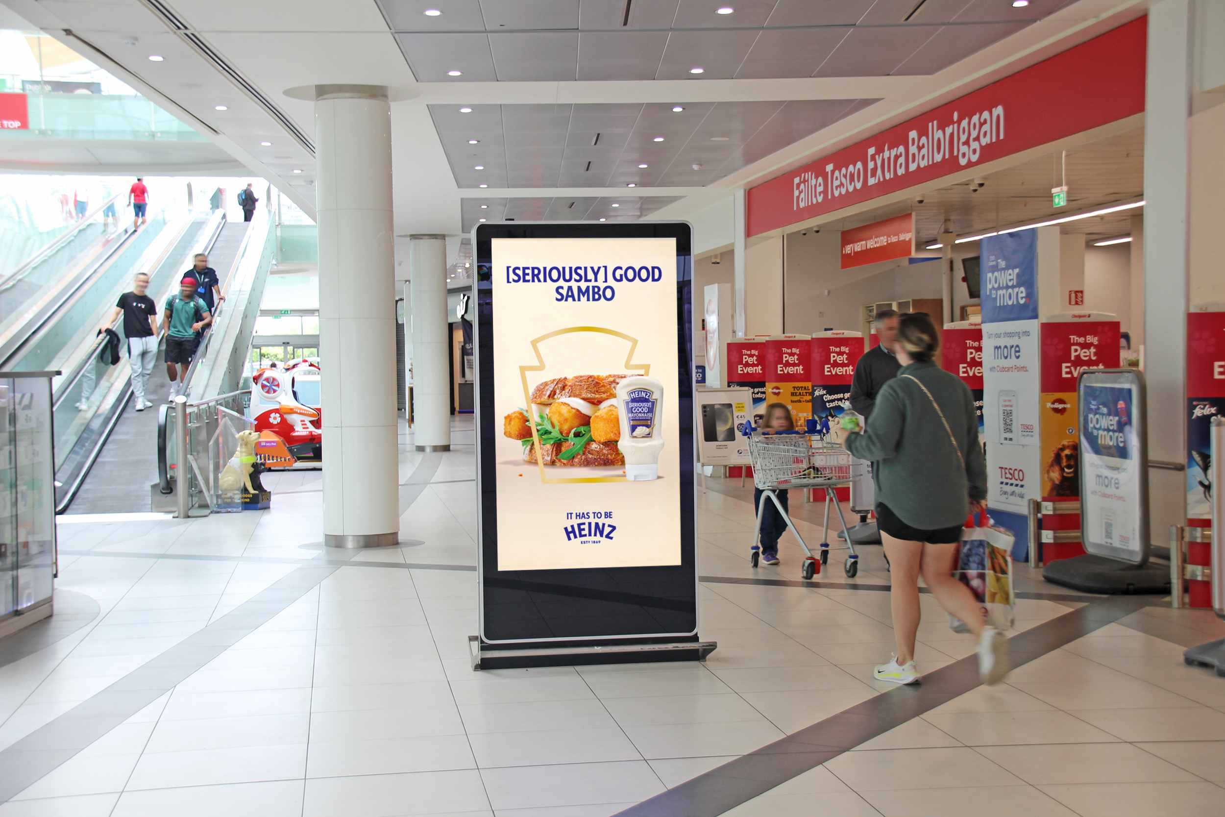 An Adshel Live Retail digital screen displaying an advertisement for Heinz Mayonnaise, situated outside the entrance of a Tesco supermarket in a shopping mall environment