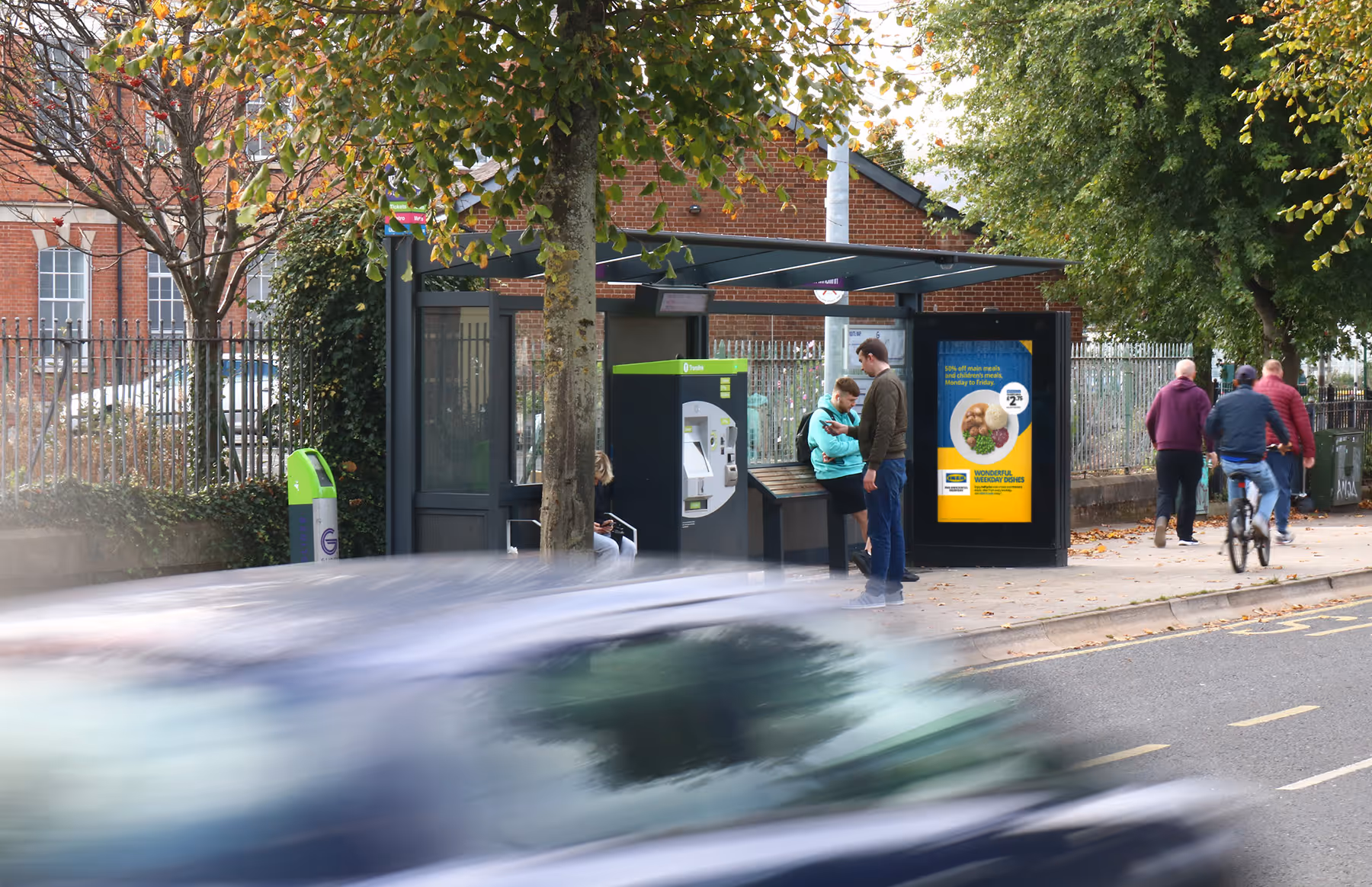 A digital bus shelter advertisement located on a busy road with people and traffic passing by