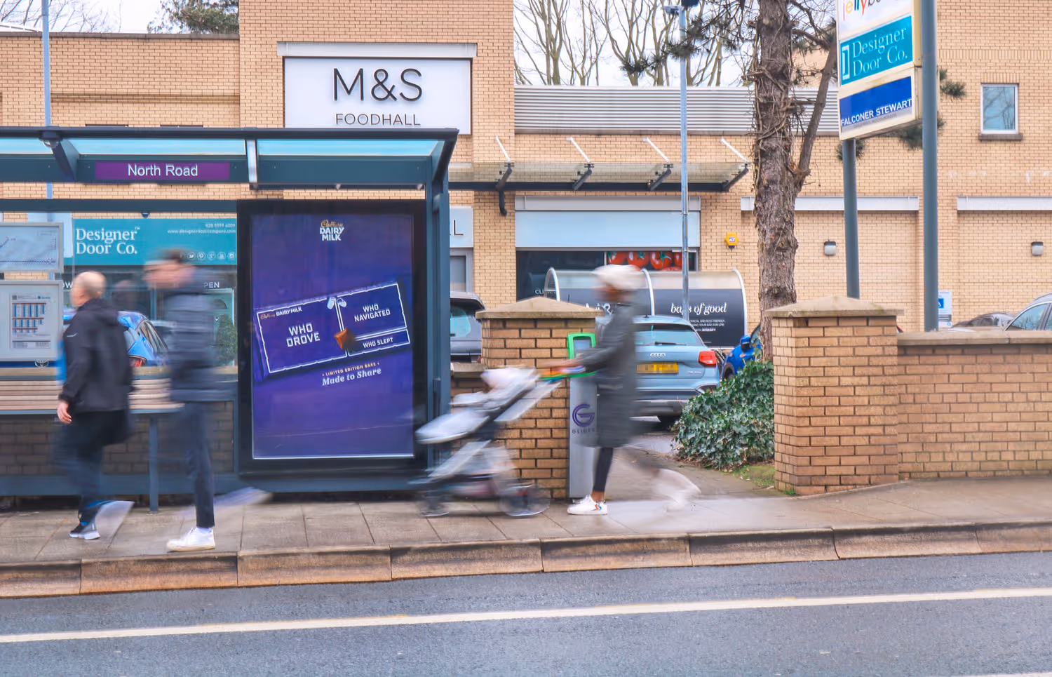 An Adshel bus shelter advertisement on a busy road with people passing by