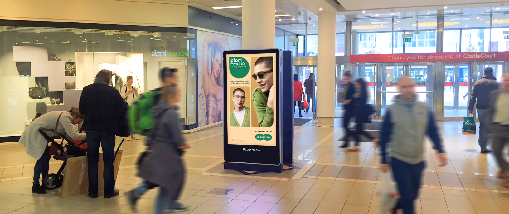 A digital mall advertising screen located at the entrance of a busy shopping mall with shoppers walking by