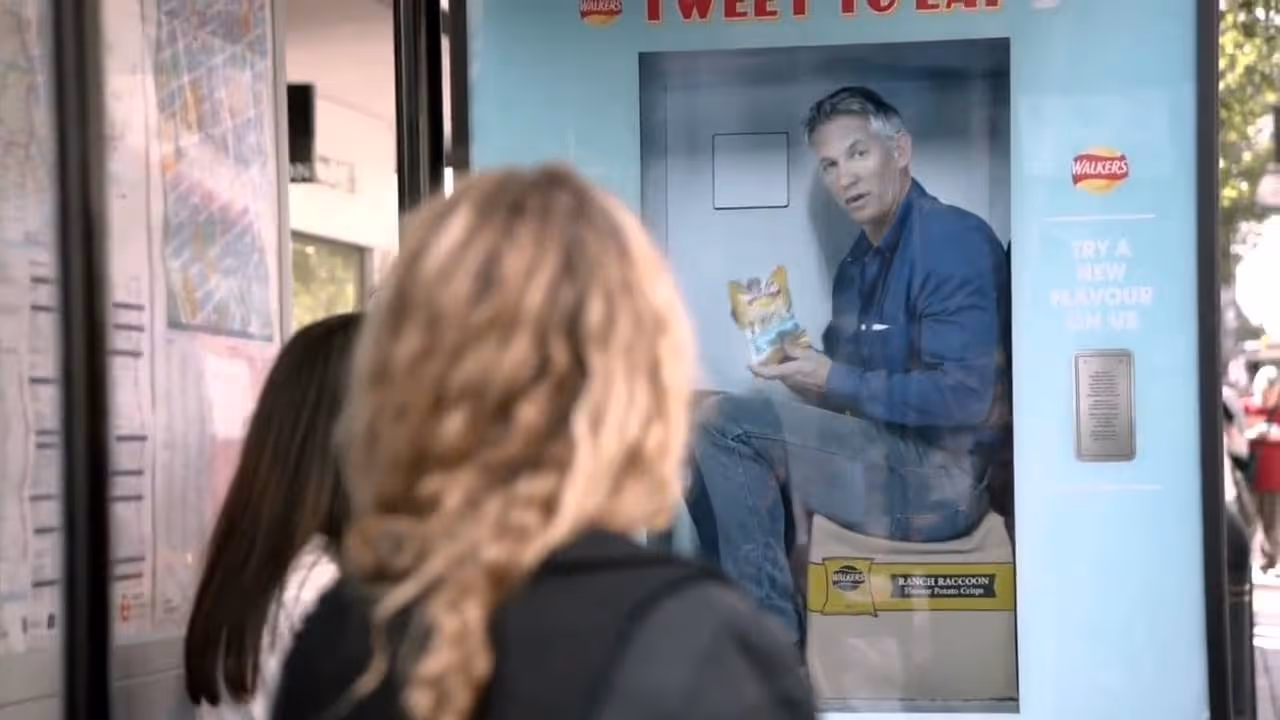 Gary Lineker on a bus stop advert holding a Walkers bag as a lady looks on.