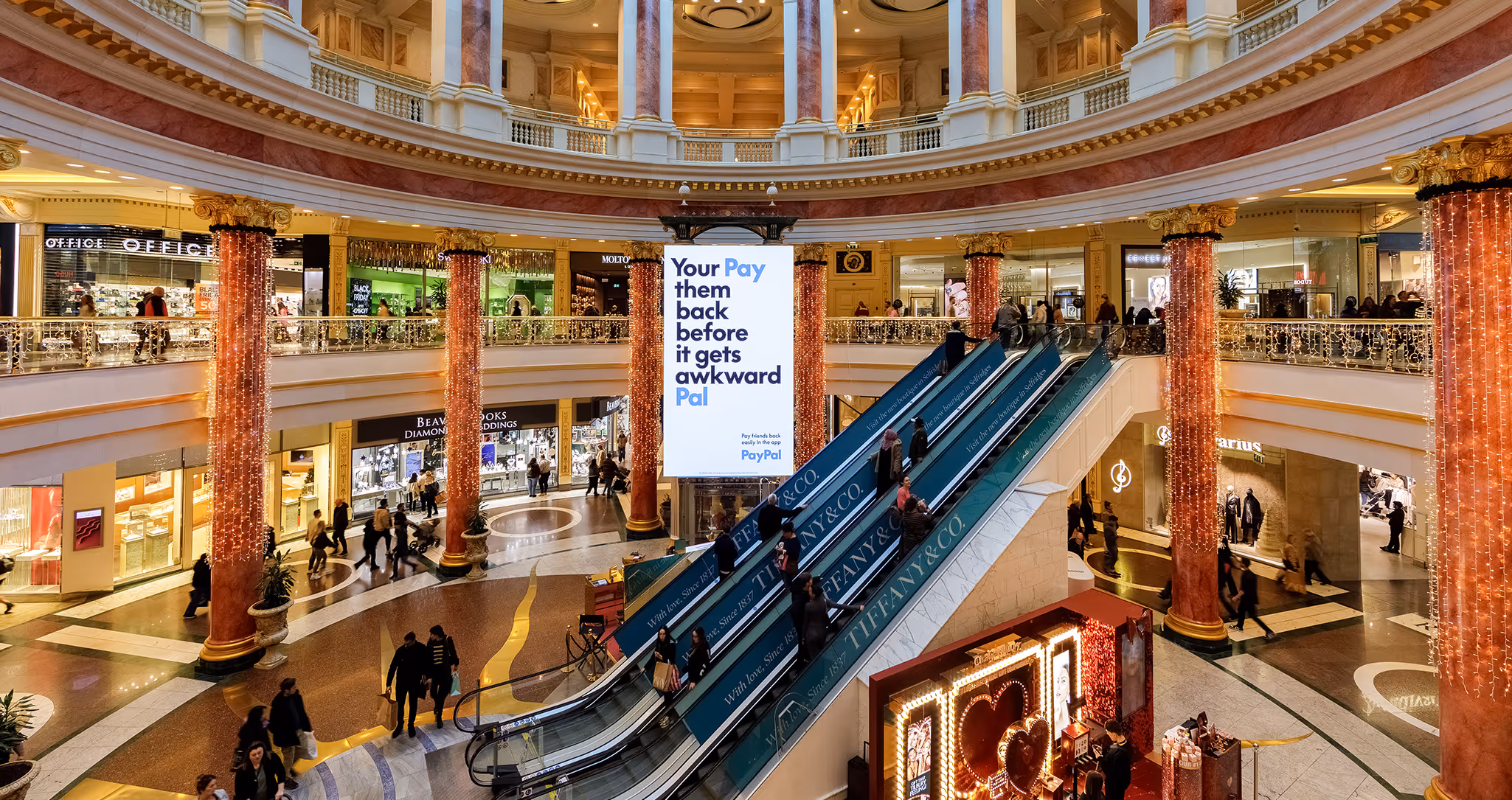 Escalators in a mall next to a very large digital outdoor advertising screen.