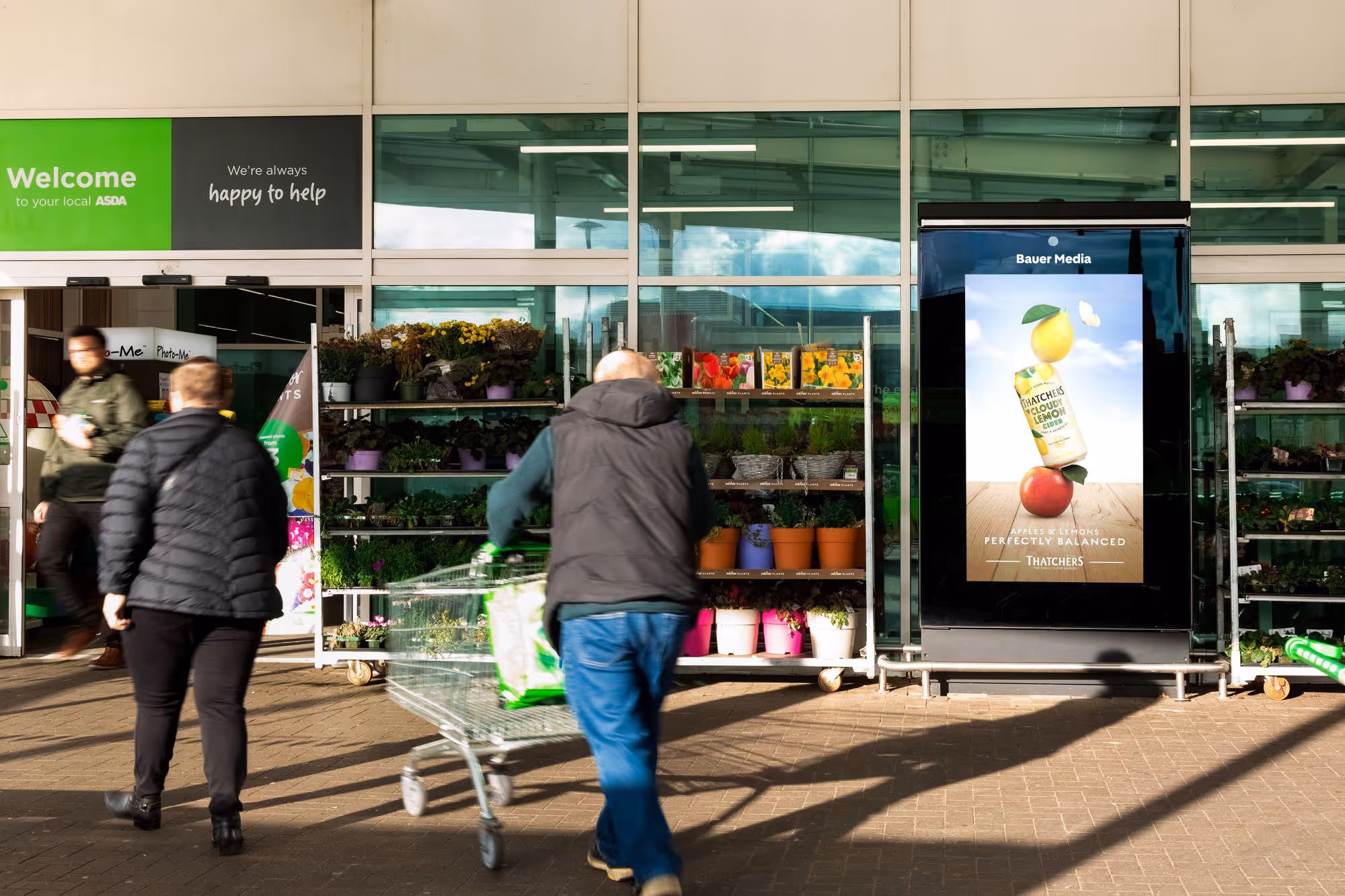 Shoppers walking into a supermarket. There's a digital screen outside next to flowers. It's an Asda store.
