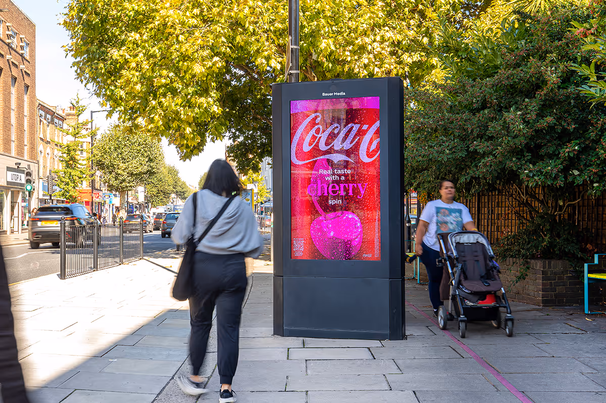 People walk past a digital screen on a summers day. The poster is for Coke Cherry.