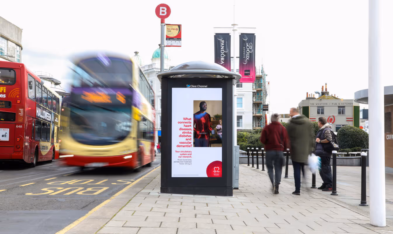 British Heart Foundation advertising on a digital screen at a bus shelter as buses and pedestrians pass by 