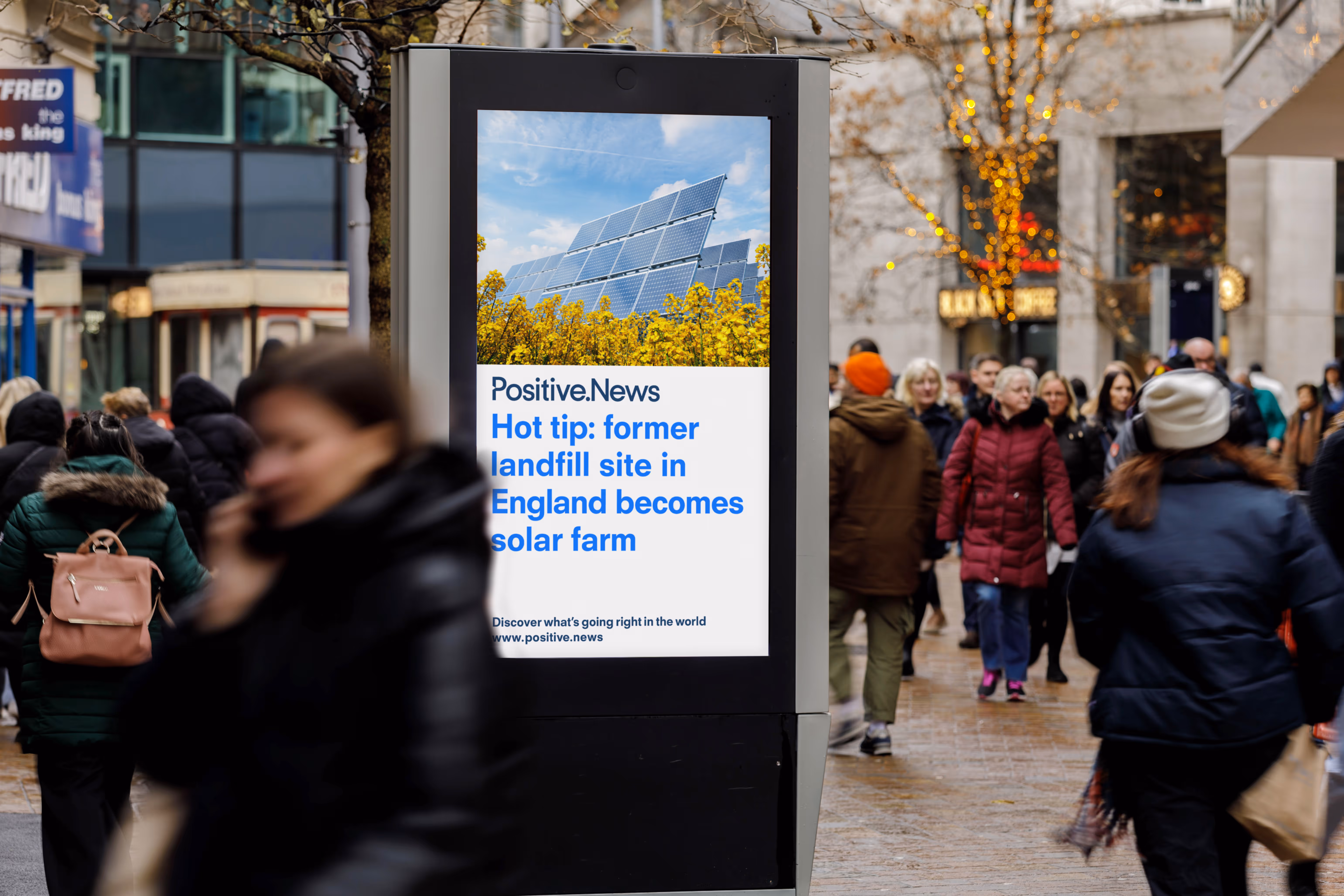 A Positive.News headline displayed on a Bauer Outdoor Media UK Adshel Live display on a busy street as people walk past during the day