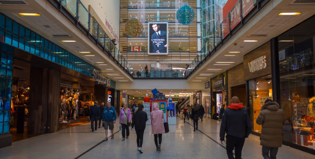 A Prada L'Homme advert features on Malls Live in a busy shopping centre during the day as shoppers walk through