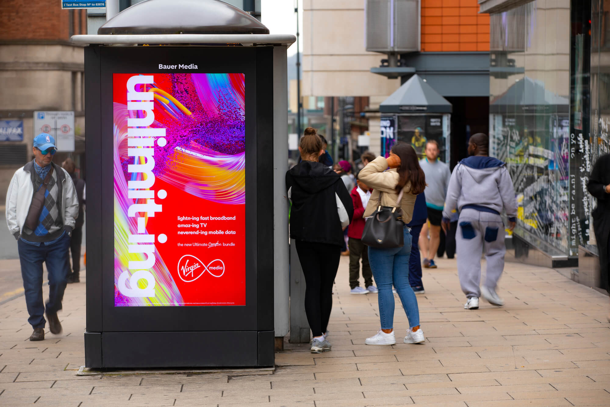 A Virgin Media advert on a digital screen, on a busy sidewalk, during the day, as pedestrians pass by 