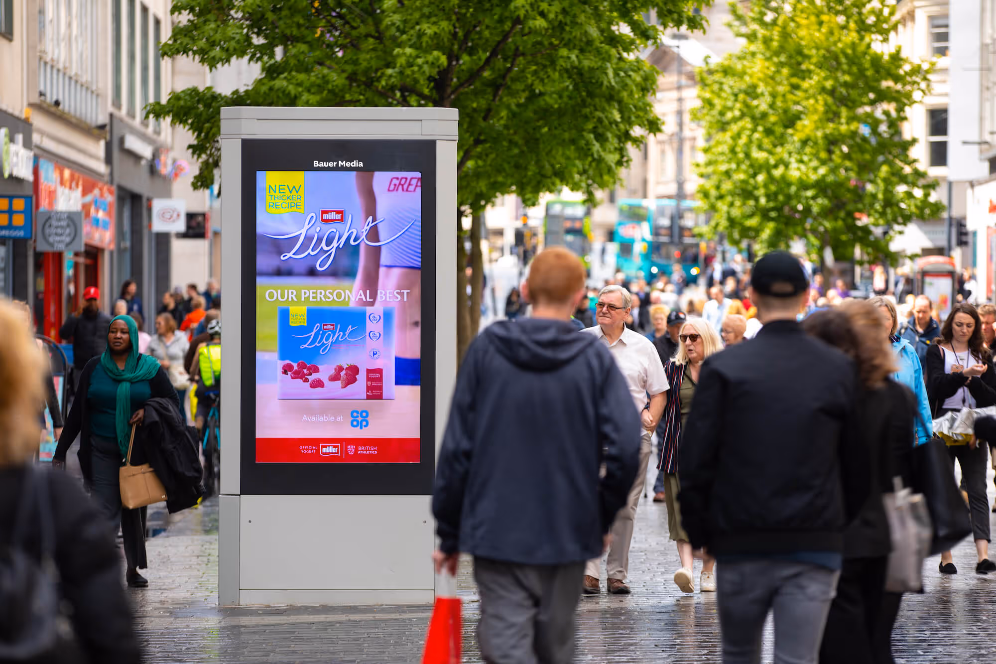 A Muller advert on a digital screen, on a busy sidewalk, during the day, as pedestrians pass by 