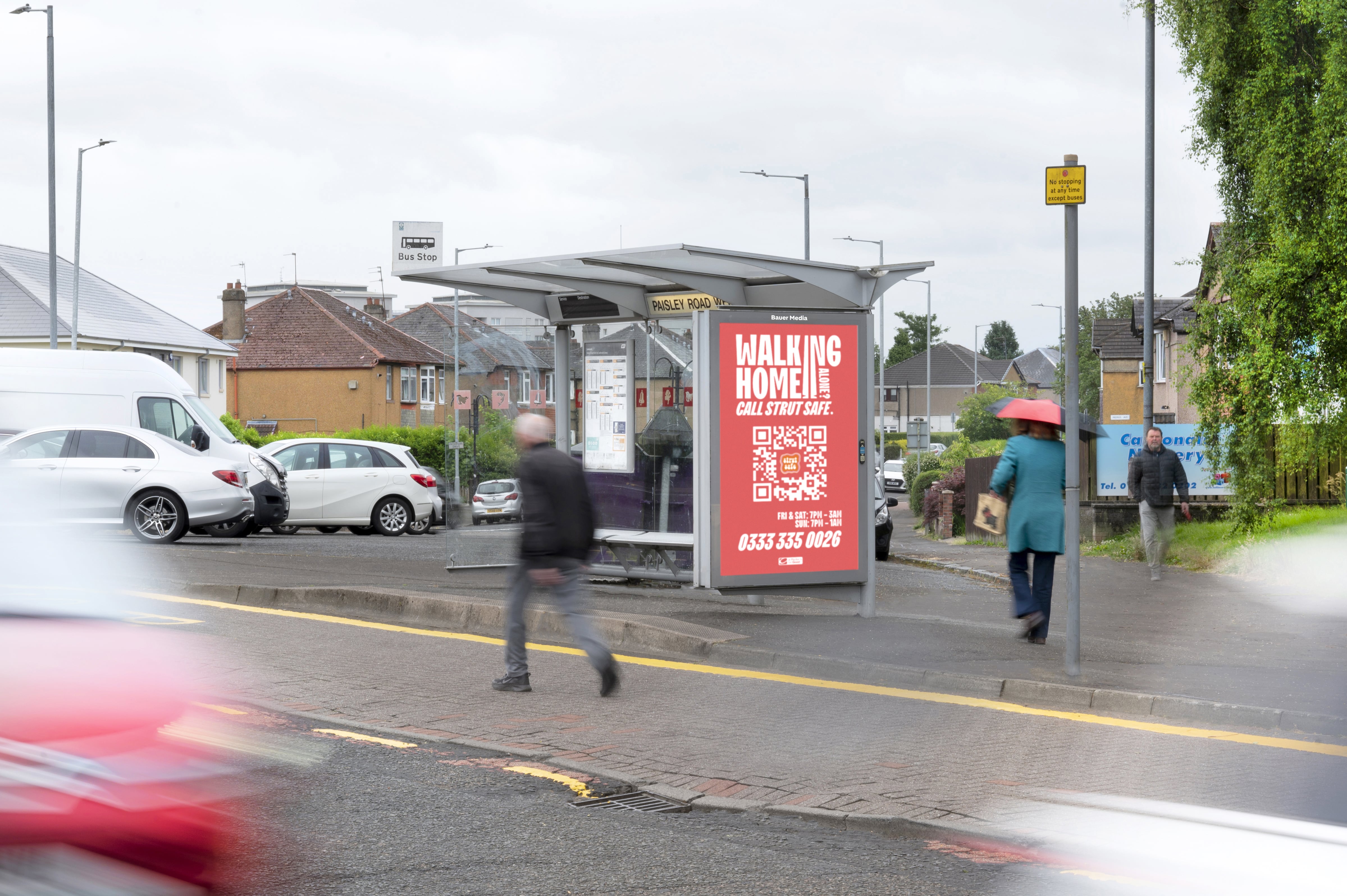 A Strut Safe QR code features on the side of a bus shelter, during the day, as cars and pedestrians pass by