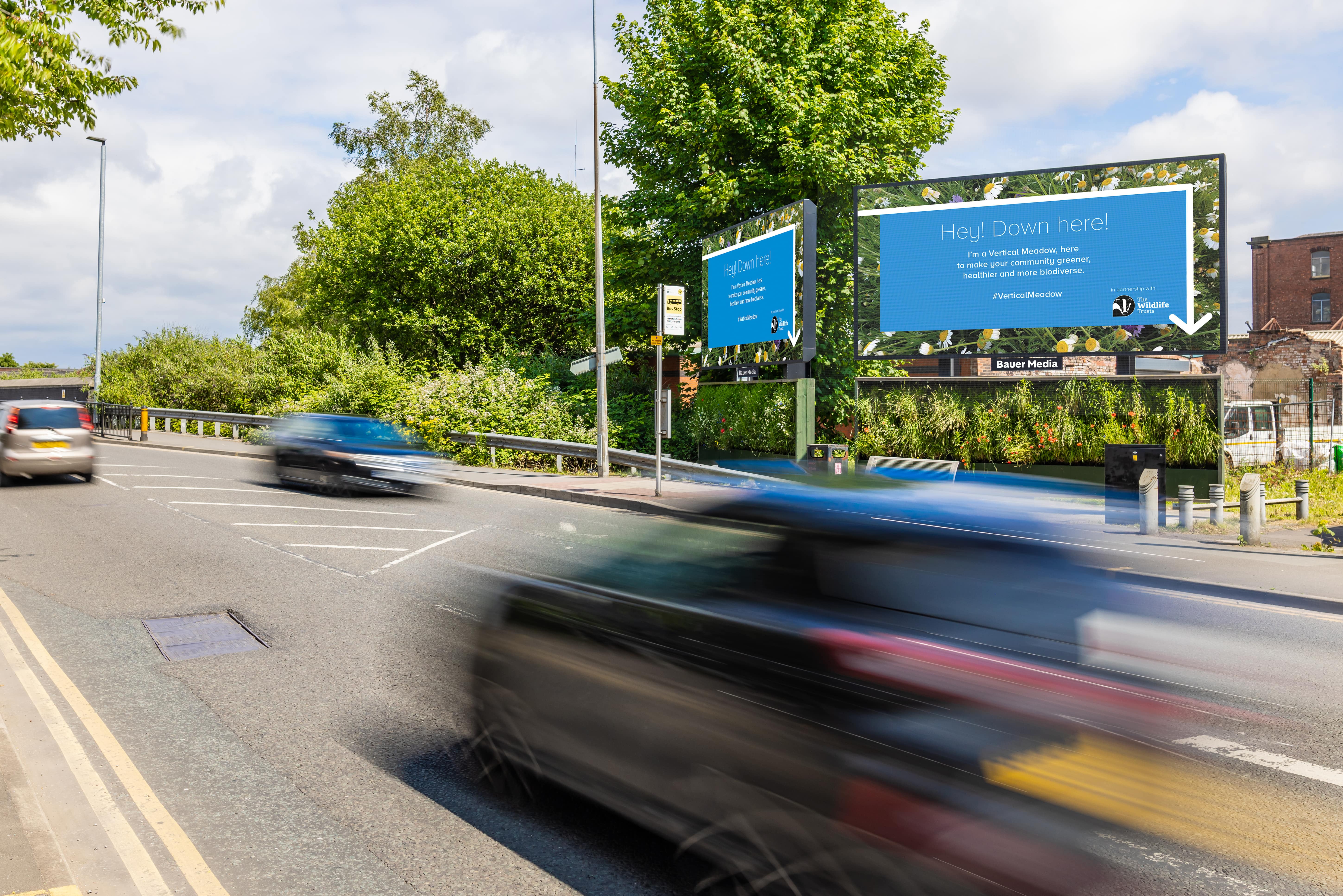 Two Vertical Meadows sit by a busy road, with cars driving by and a billboard overhead featuring an advert for Vertical Meadow that says "Hey! Down here!"