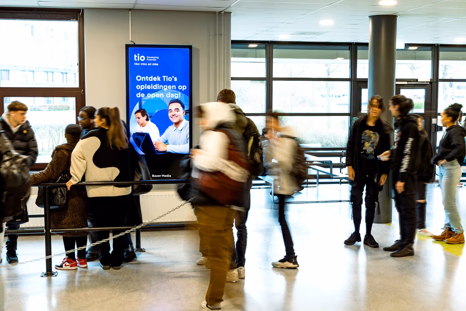 Digitaal scherm hangt aan wand in kantine en scholieren lopen voorbij of staan stil