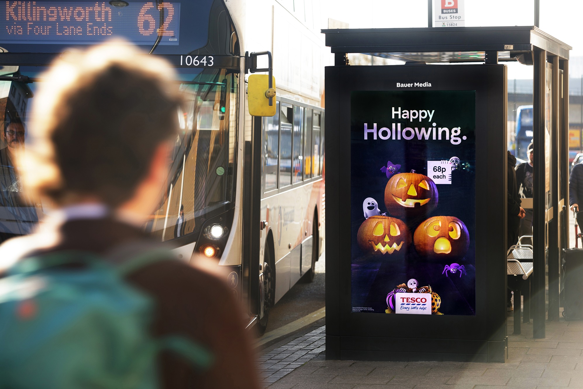 Tesco Halloween advert on a digital screen, on the side of a bus shelter, on a busy street as buses and pedestrians pass by during the day