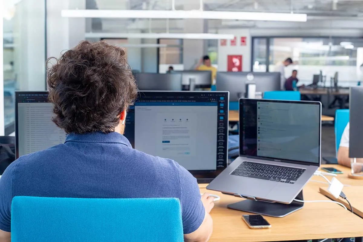 A man is working at a desk with two monitors and a laptop in a modern open office, focused on his screen.