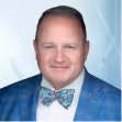 Middle-aged man wearing a blue checkered blazer and a colorful bow tie, smiling against a light blue background.