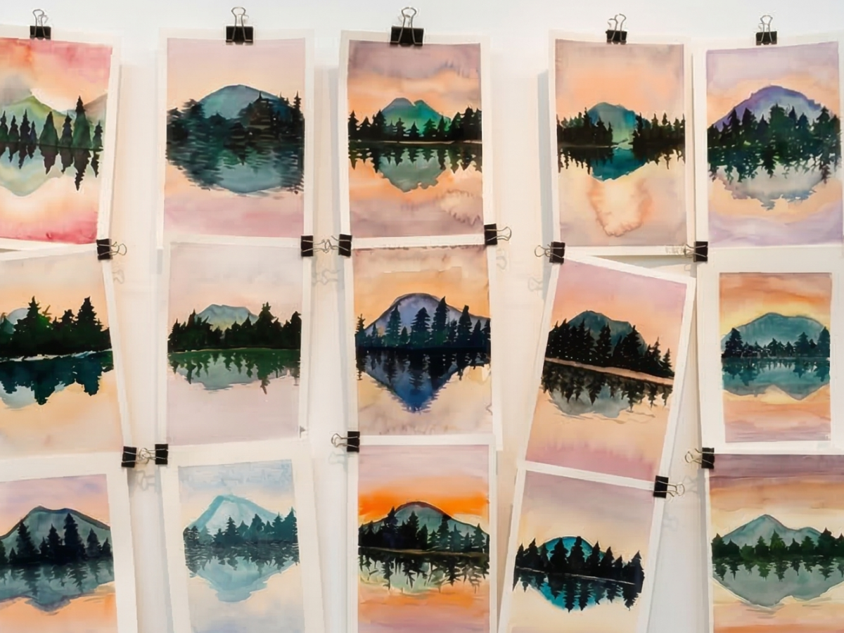 Une personne dessine avec un stylo fin sur une grande feuille blanche avec des taches colorées à l'aquarelle, à côté d'une palette de peinture et d'un carnet jaune.