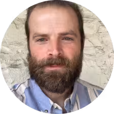 Bearded man with short brown hair wearing a light blue collared shirt standing in front of a light stone wall.
