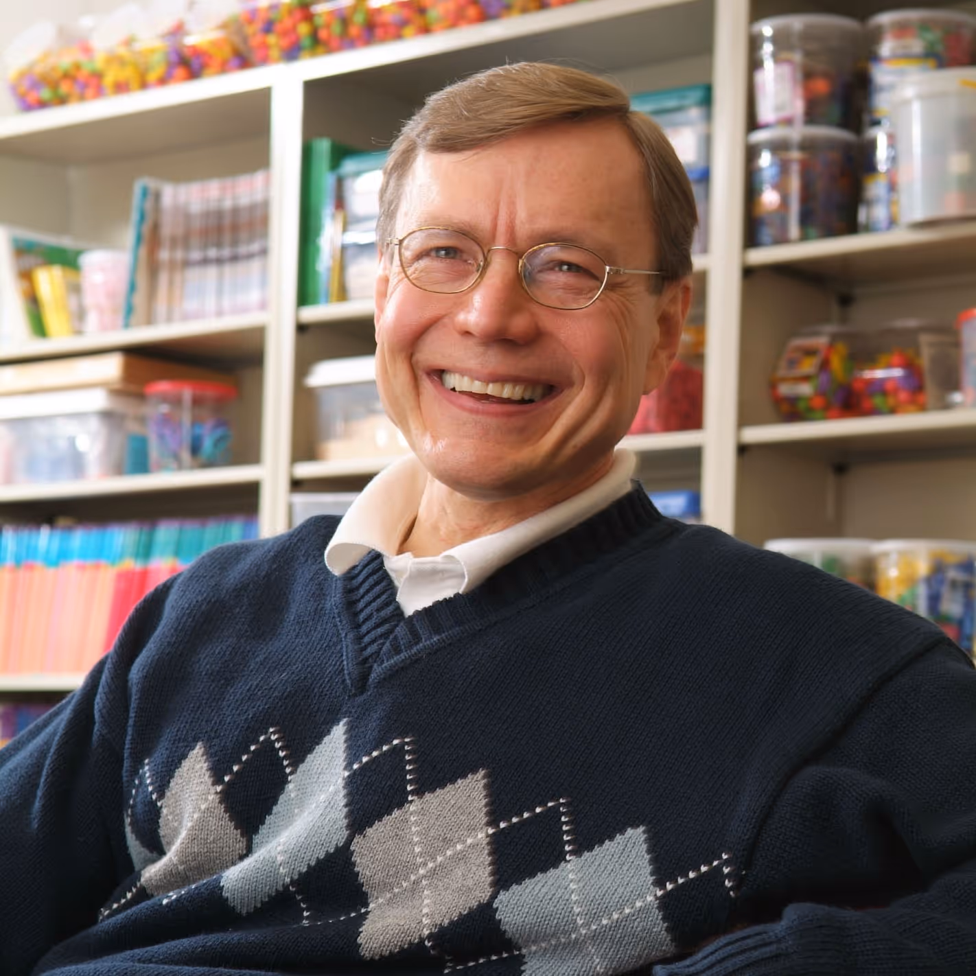 Smiling middle-aged man wearing glasses and a navy blue argyle sweater in a room with shelves holding colorful educational materials and containers.