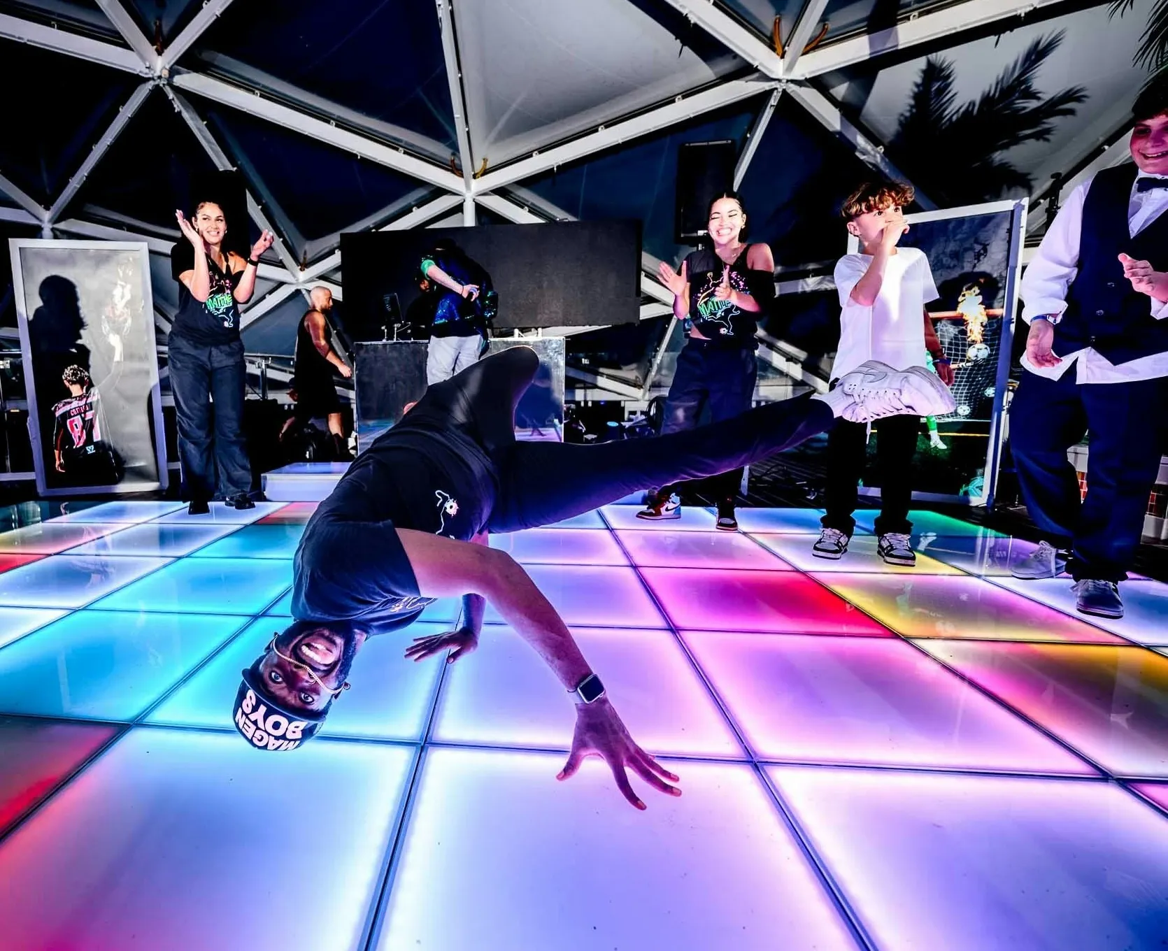 Man breakdancing on a colorful illuminated floor while a group of people cheer him on in a tent-like structure.